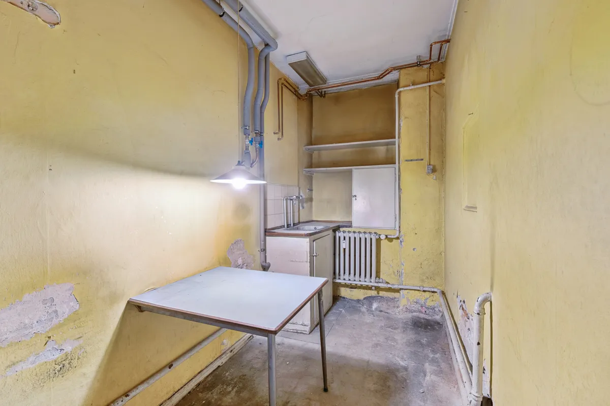 En stor lys stue med gammelt gulv og lysegul væg. Et hængende lysekron, et hvidt bord og et køkken med radiator og loftslampe.