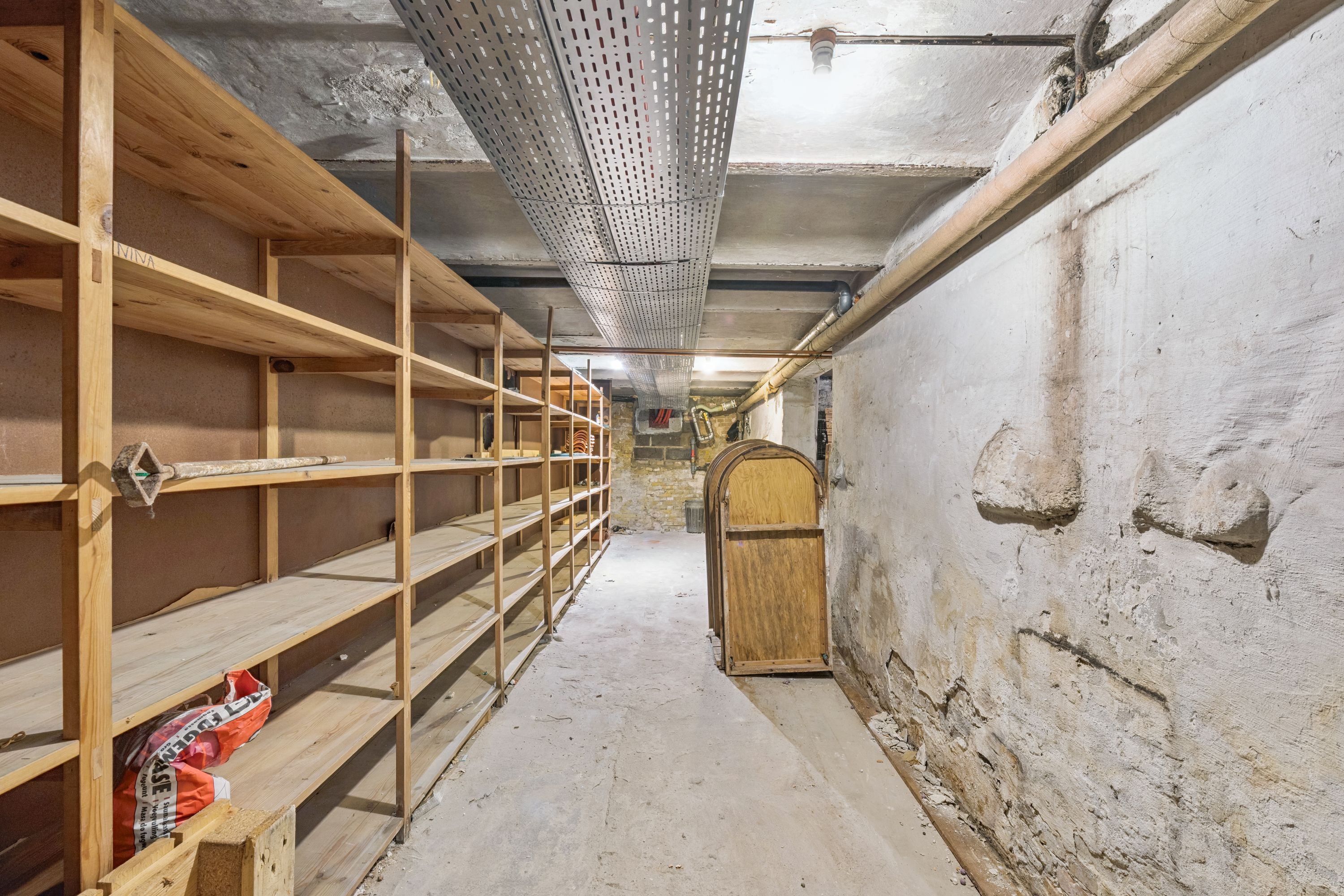 Lejlighed med lys kælder og træhylder, røgskader på væggen, sammen med rør og kabelkanaler. Forskellige genstande er placeret på hylderne og et stel er fastgjort til væggen.