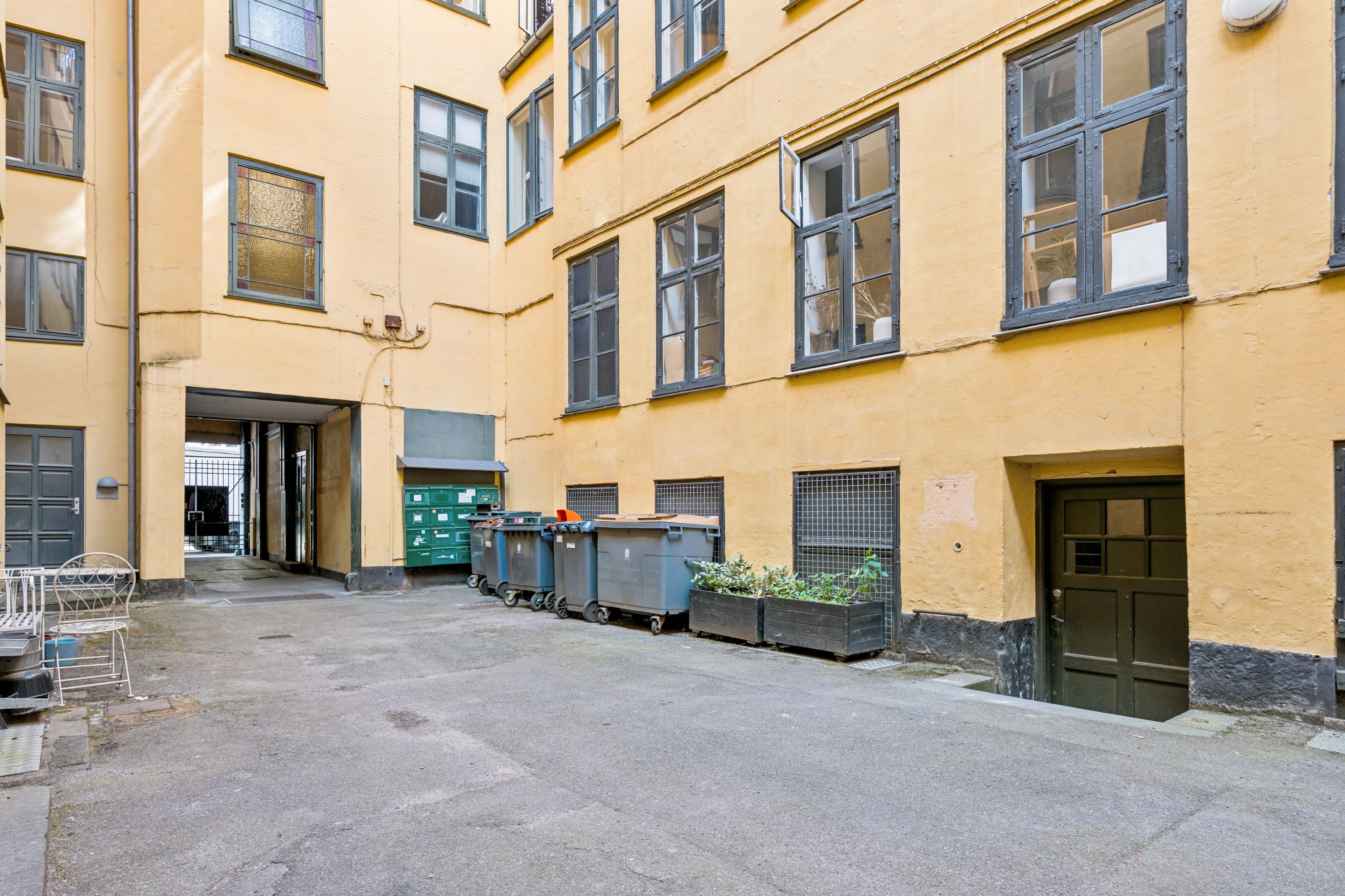 Lyst og indbydende stueplan med to plantekasser og affaldscontainere samt to adgangsdøre i en gul bygning.