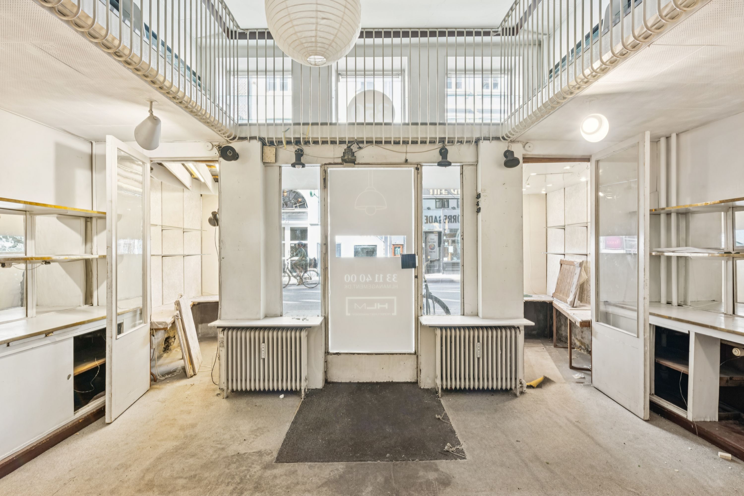 En tom kontorlokalitet med åbne døre og lyse vinduer, radiatorer og et hængende lys. Den har hvide vægge og lofter samt et indgangsparti med mørke gulvbelægning