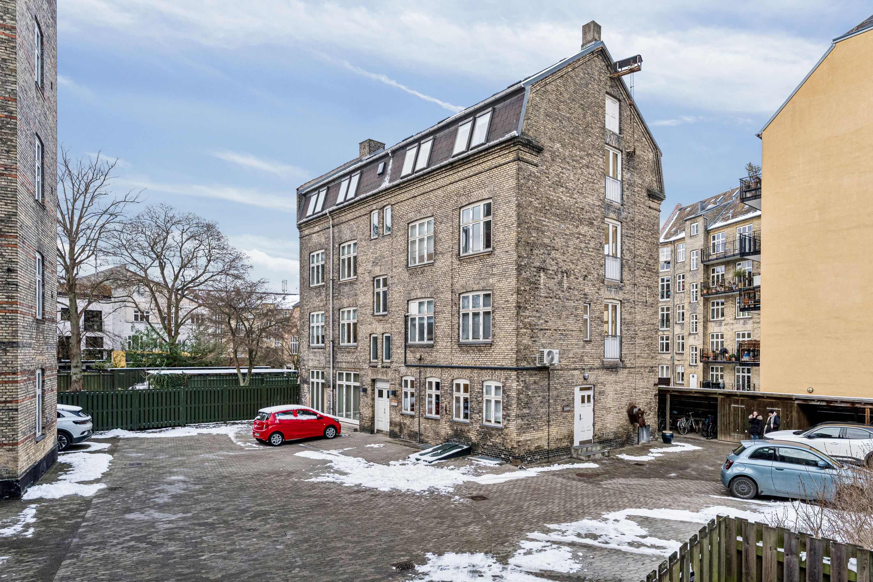 Kontor til leje på Smallegade 46B i Frederiksberg med en stor guldbrikket bygning, to biler parkeret og mennesker i baggrunden. Bygningen har flere vinduer og en hvid dør.