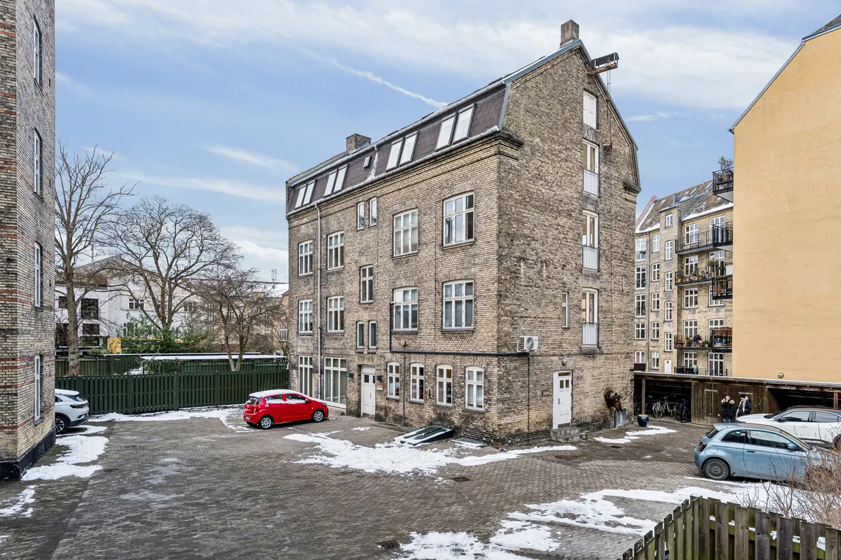 Kontor til leje på Smallegade 46B i Frederiksberg med en stor guldbrikket bygning, to biler parkeret og mennesker i baggrunden. Bygningen har flere vinduer og en hvid dør.