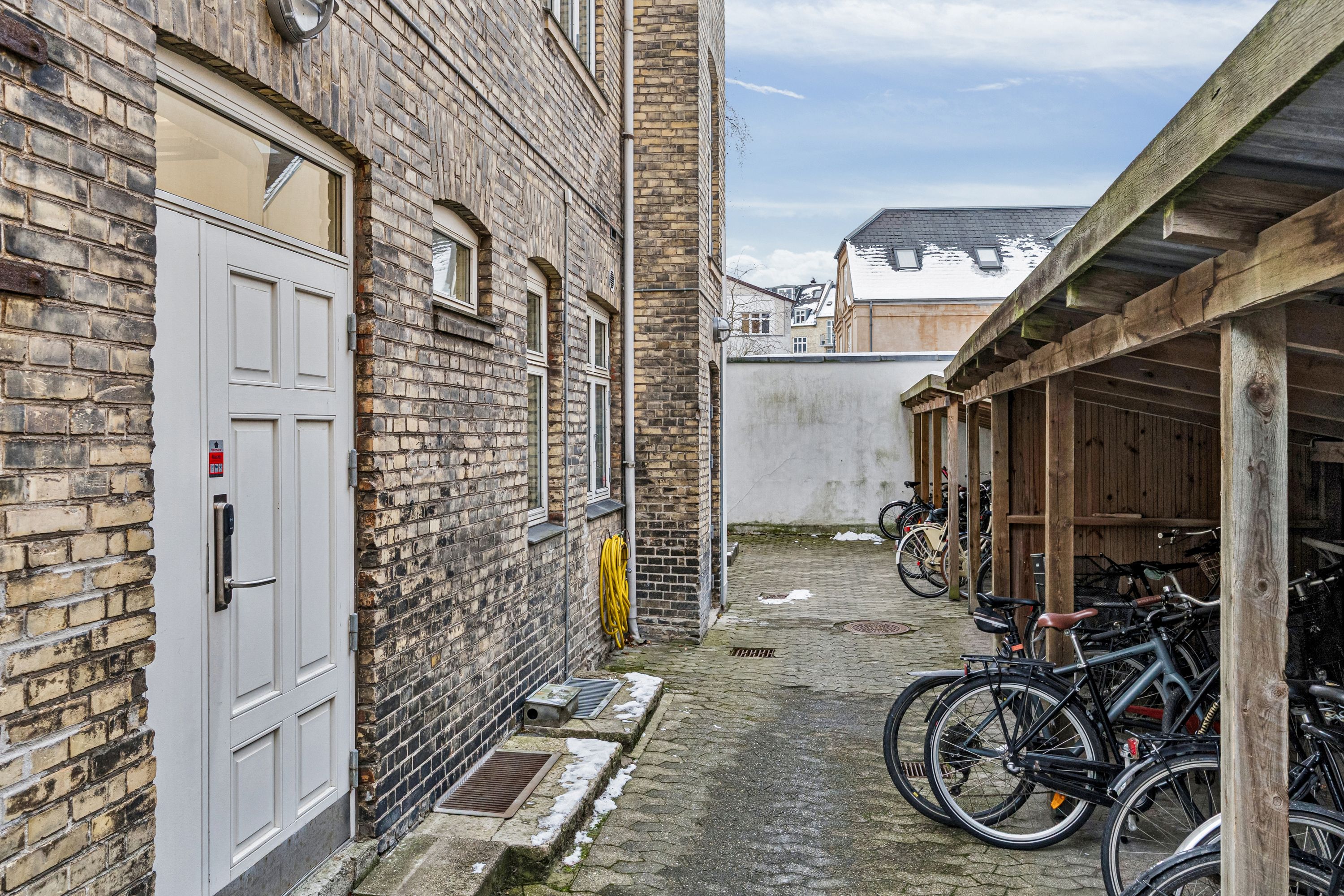 Garage med to cykler parkeret og et stort hvide dør i baggård med træhytte og stående cykler