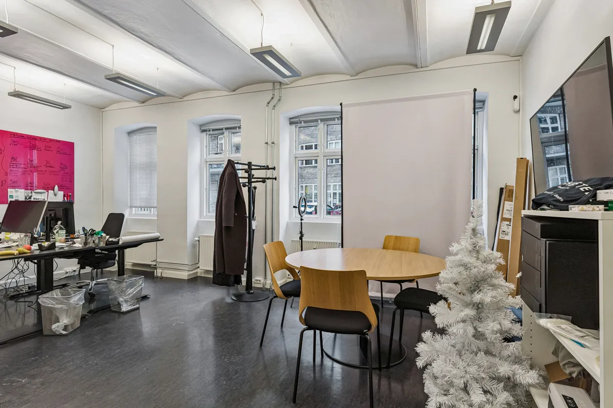 Kontor med rundt bord og stole, en pink tavle og en hvid juletræ samt en skrivebord med computer og skraldespand.