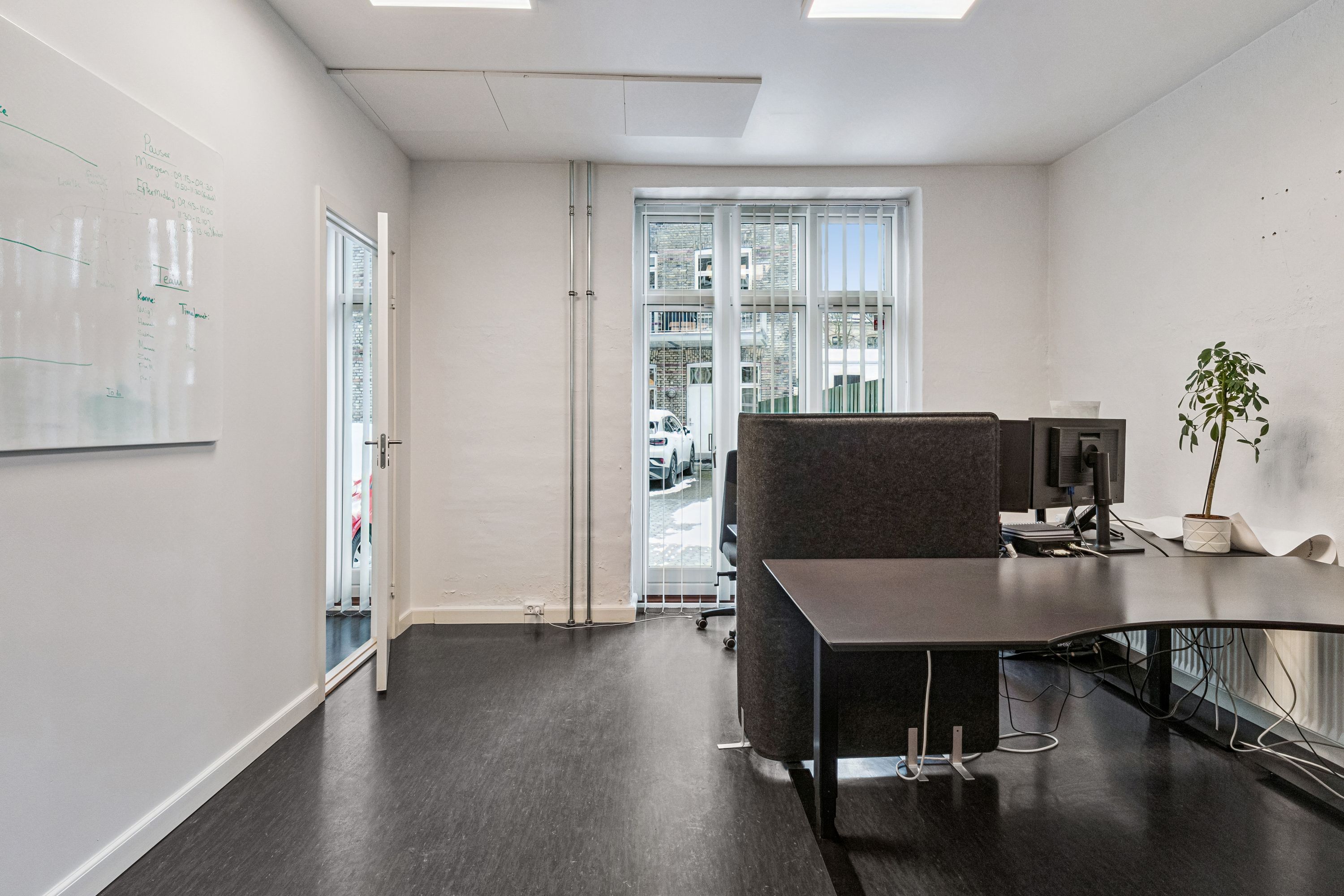 Kontorrum med højloft og hårdt gulv, hvide vægge, moderne møbler, plante og skrivebord med computer og skærm samt åben dør med udsigt til gårdsplads og biler