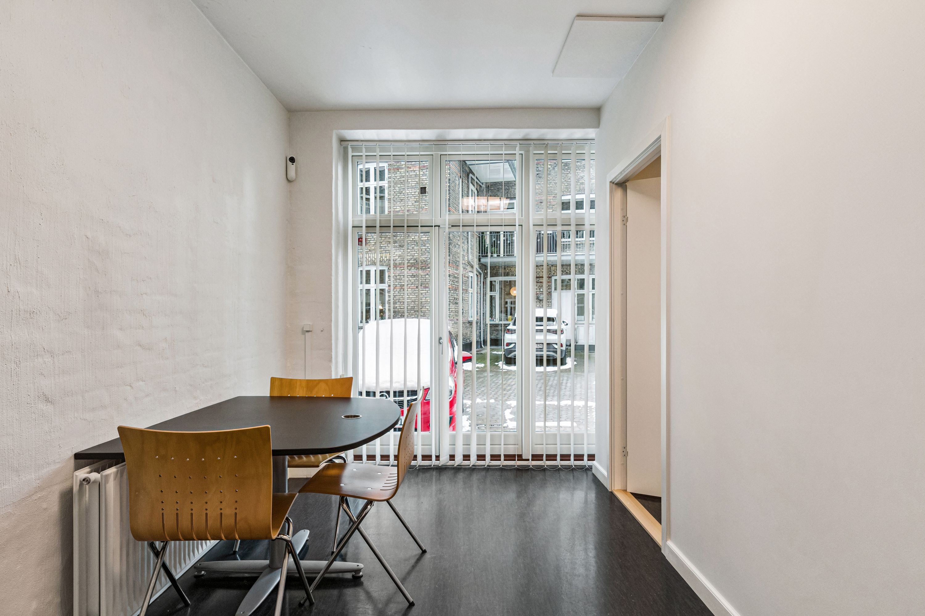 Kontor med møbelsæt og højt vindue med persienner mod gaden. Kontoret har en rund bord og fire stole. Der er et radiator monteret på væggen.