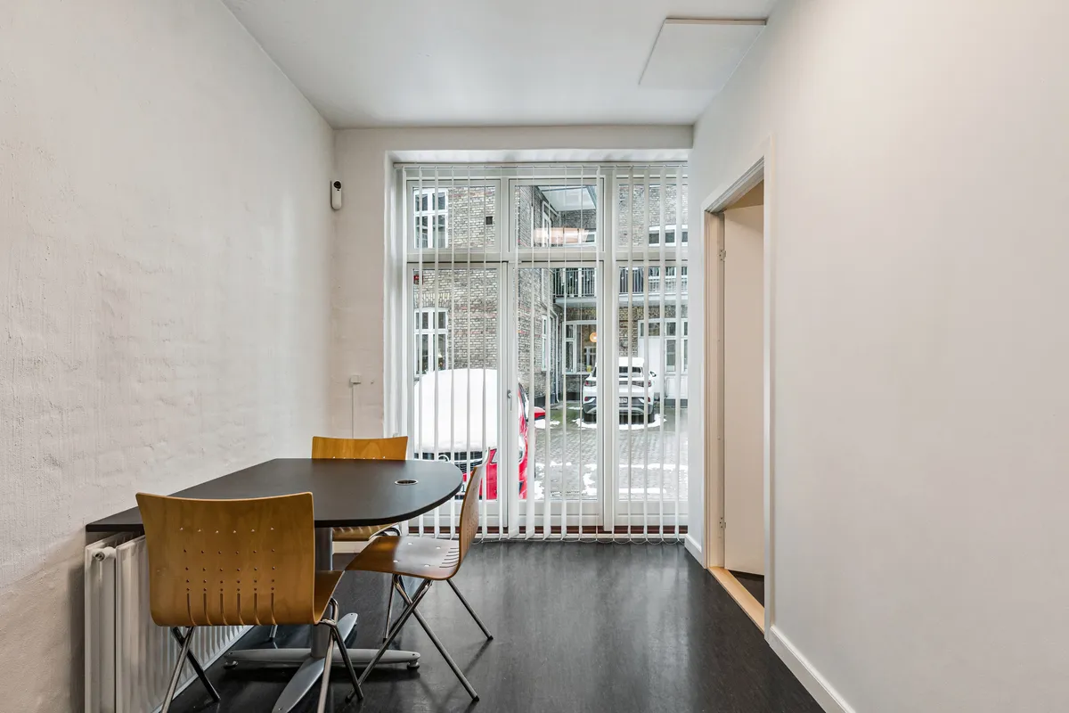 Kontor med møbelsæt og højt vindue med persienner mod gaden. Kontoret har en rund bord og fire stole. Der er et radiator monteret på væggen.