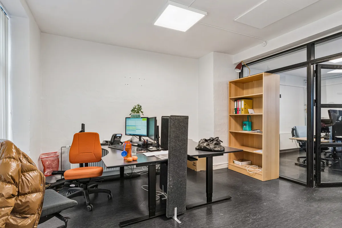 Kontor med computer, monitorer og skrivebord. Sofa med skindfrakke og rødt stolpebord. Skrivebord med telefon, skraldkurv og plante. Højt hylde med papirer og taske.