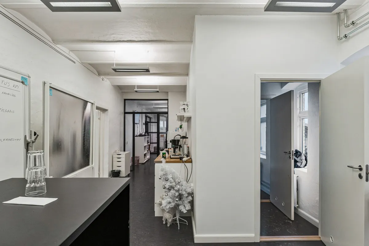 Kontor med lyse loftsvinduer, hvide vægge, en sort bord med glas og papir, en juletræ med hvidt sne og lysereflekser, samt en åben dør med et skur og et vindue.