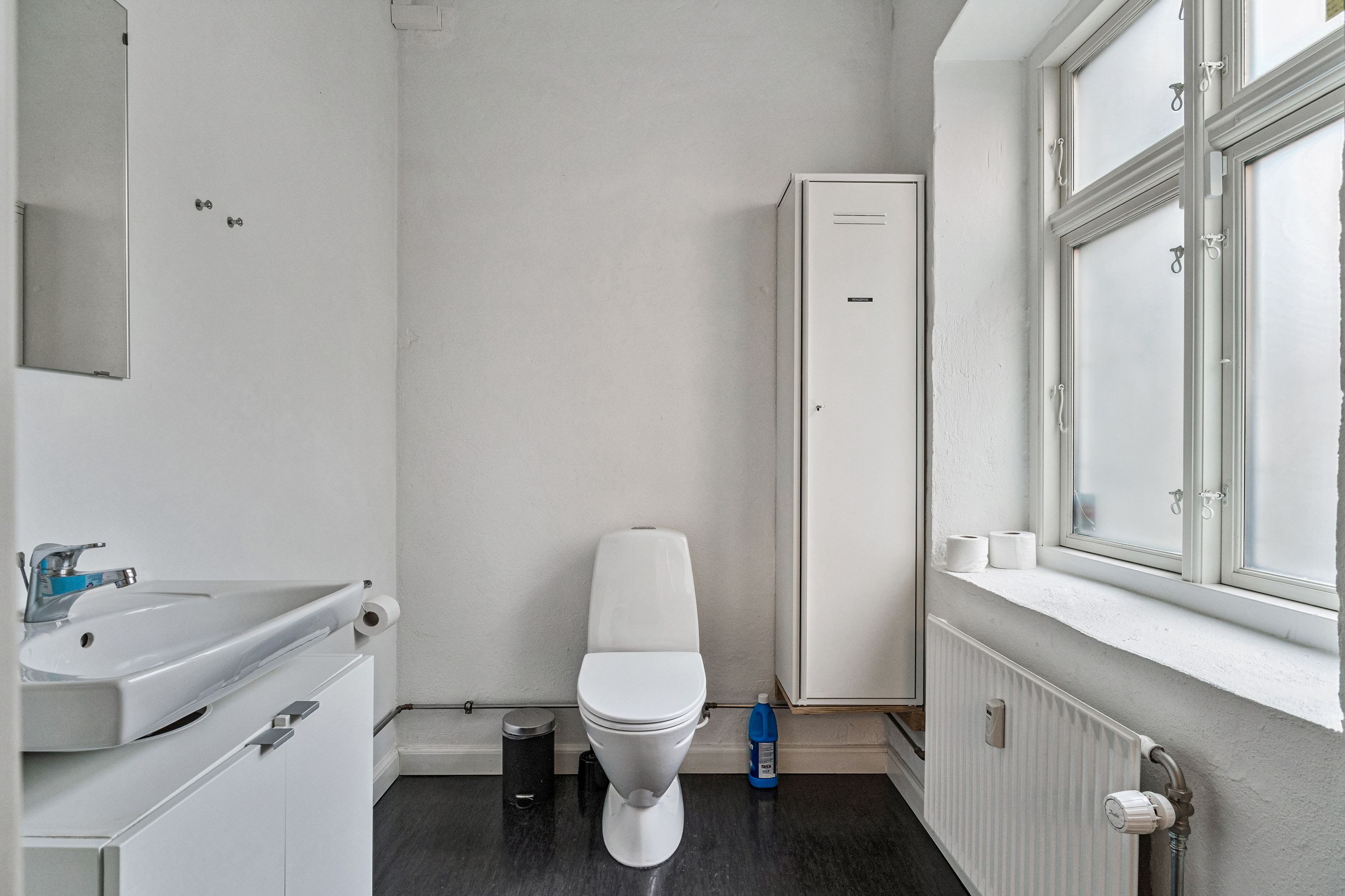 Kontor på Smallegade 46B med hvid toilet, radiator, strygejern og strygebræt, samt hygiejneprodukter og sæbe.