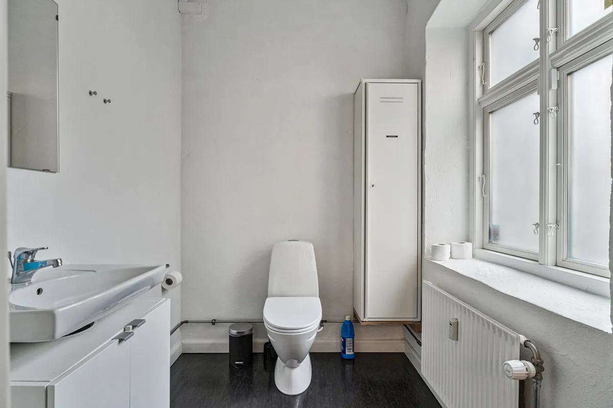 Kontor på Smallegade 46B med hvid toilet, radiator, strygejern og strygebræt, samt hygiejneprodukter og sæbe.