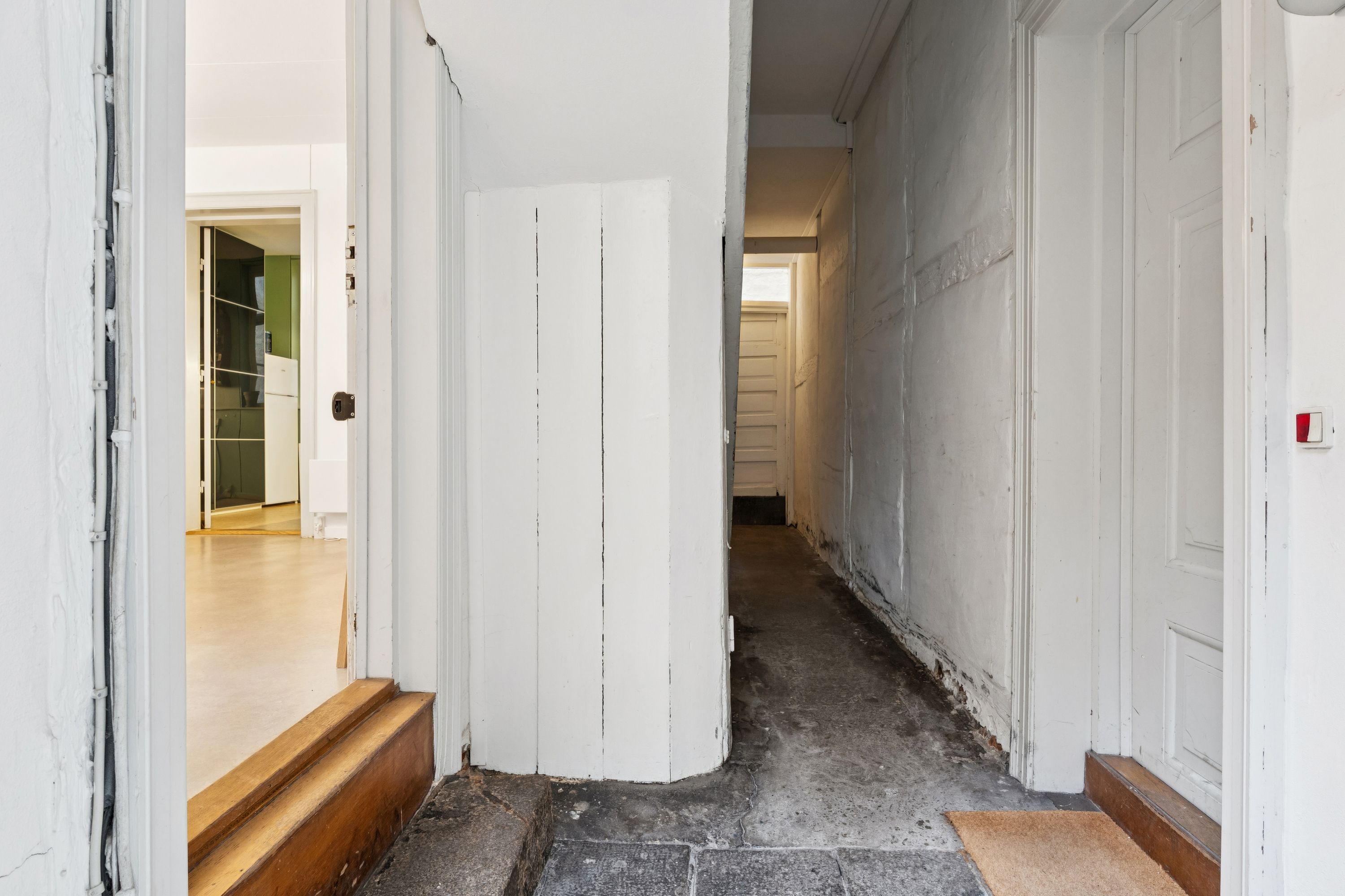 En hvid malet korridor med to hvide døre, en trætrin og en gulvbelægning, som ser ud til at være træ. Der er også en åben dør, der viser et værelse med gulvbelægning.