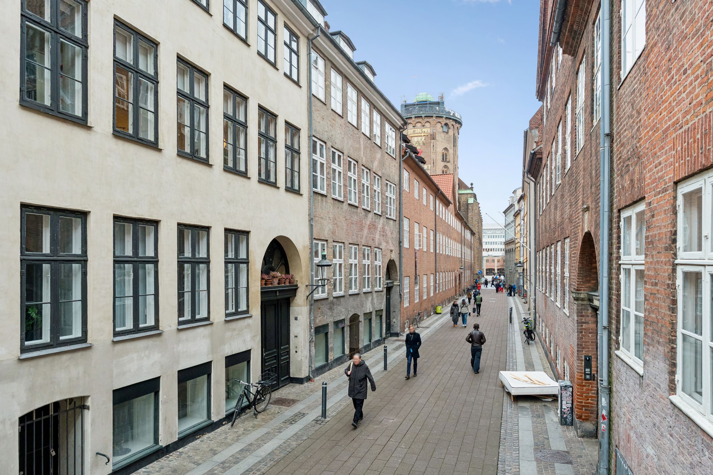 Mennesker går ad en lille gade mellem to bygninger med røde mursten og hvide vinduer. Der er et rødt hus med røde tag på hjørnet. Der er også en skulptur og et tårn i baggrunden.