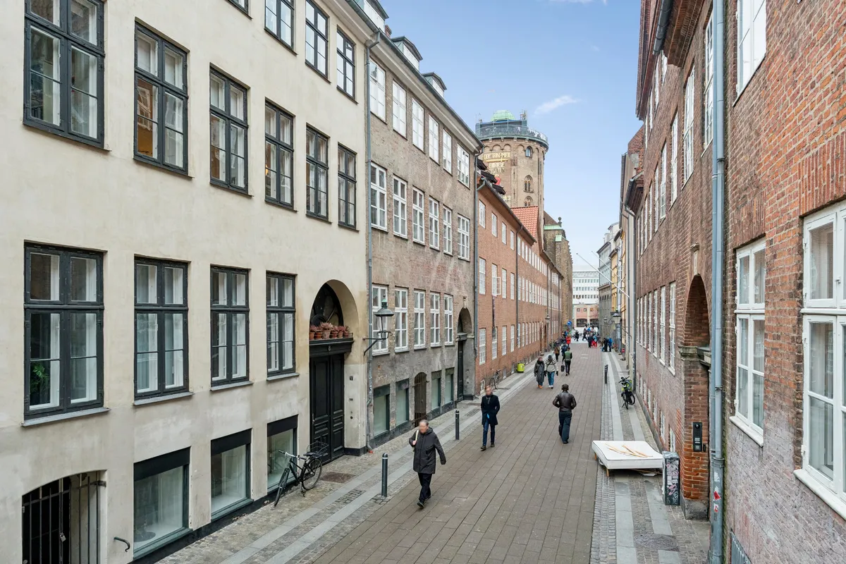 Mennesker går ad en lille gade mellem to bygninger med røde mursten og hvide vinduer. Der er et rødt hus med røde tag på hjørnet. Der er også en skulptur og et tårn i baggrunden.