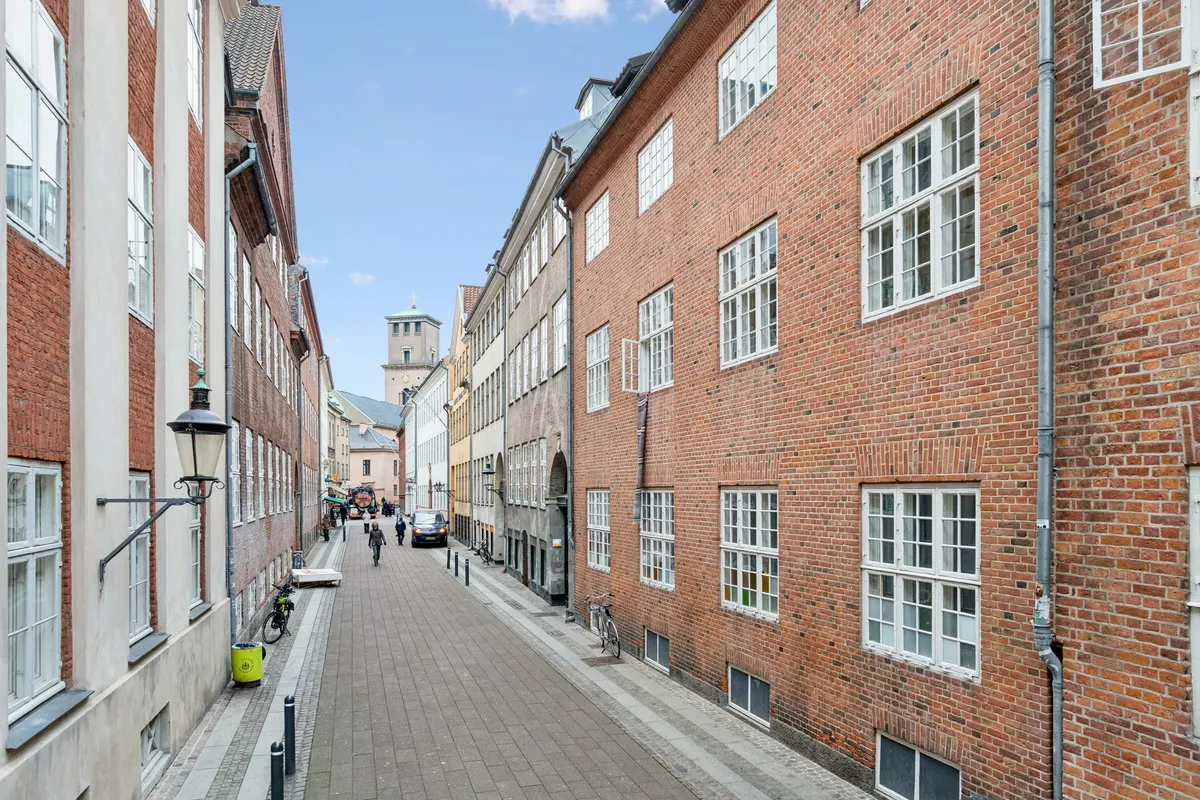 En række røde murstensbygninger og glasruder med lysegardiner i Store Kannikestræde, København K, og mennesker og biler på gaden i baggrunden