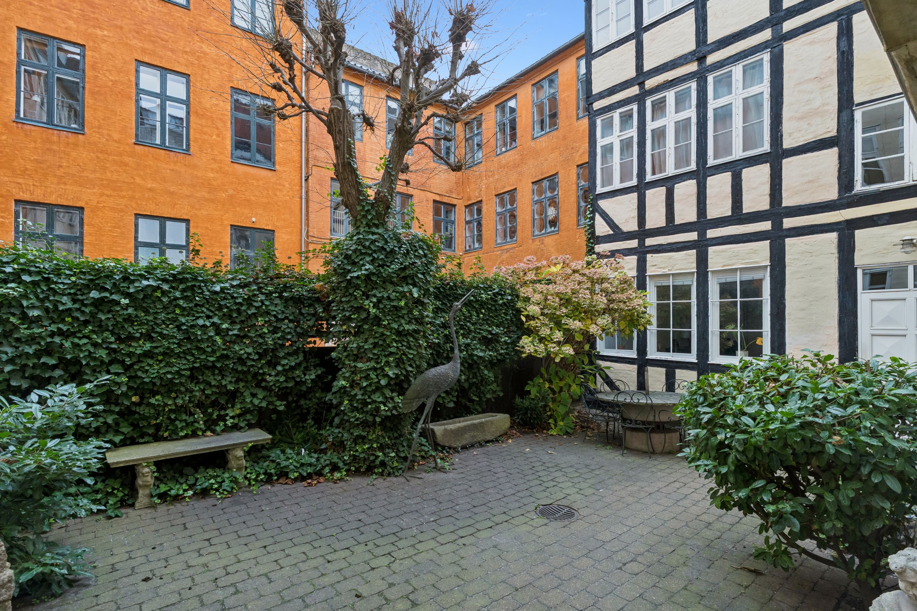 En gårdhave med to bygninger og en statue af en stork, en bænk, en lille stenbrønd og en rund bordsætning med stole og bord.