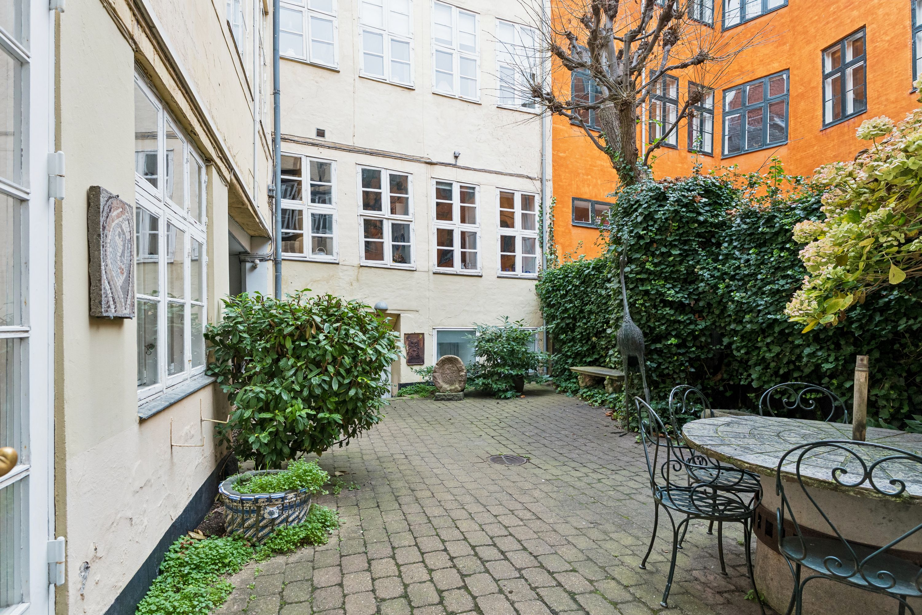 En lille have med terrasse, stole og borde, omgivet af bygninger, fuld af planter og grønne buske