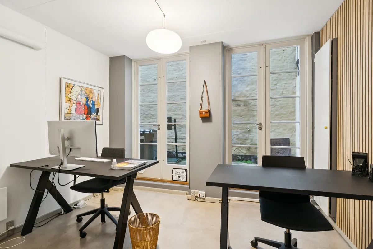 To moderne kontormøbler med computere, stole og et gulvkurv i et kontorlokal med hvide vægge og store vinduer med udsigt til en bakke