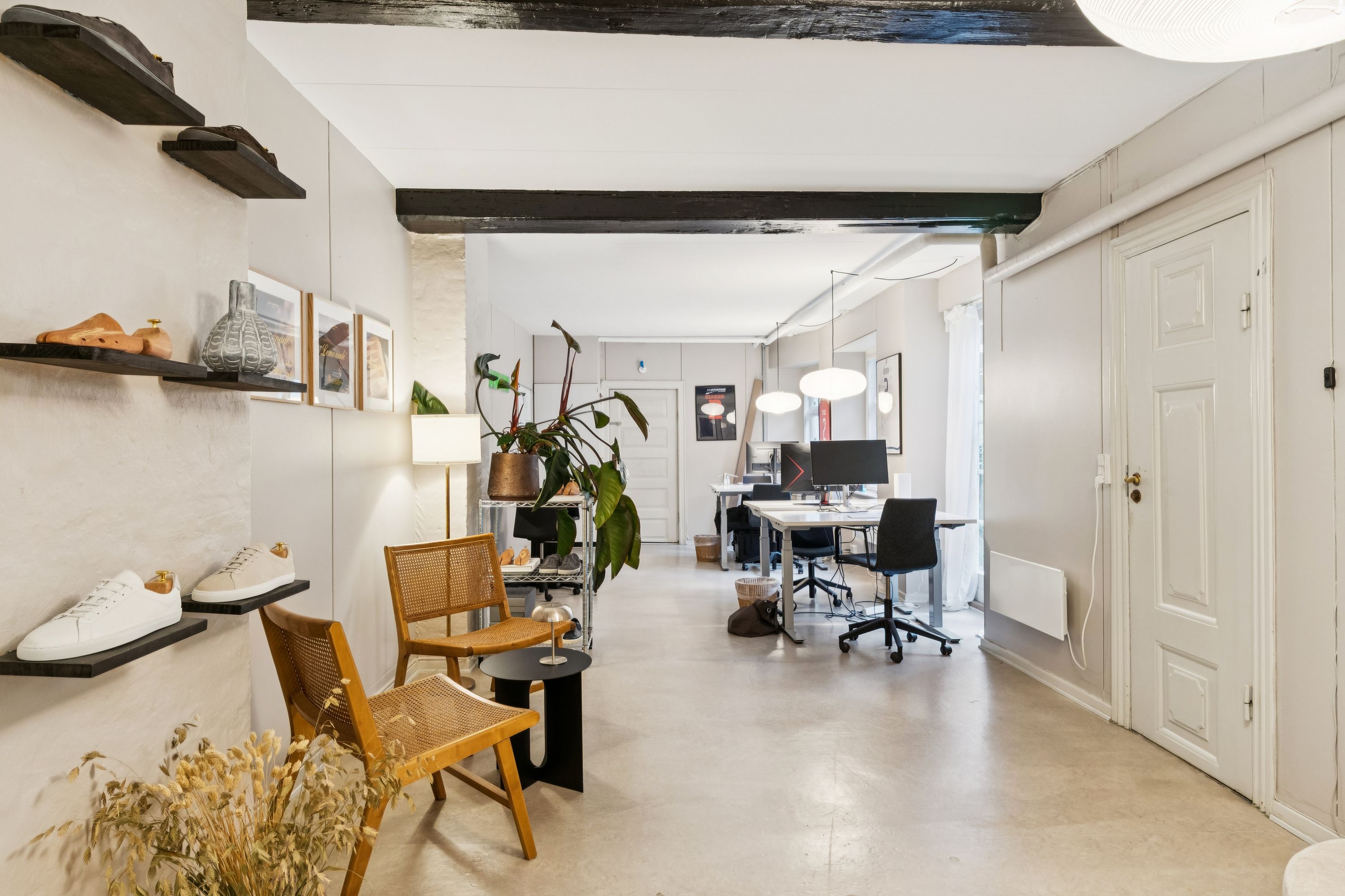 Kontor med hvide vægge, trægulv, højhængende loftsbjælker, tre skohylder med sko, tre stole, to skriveborde, en lampe, en vase, en plante og to stående lamper