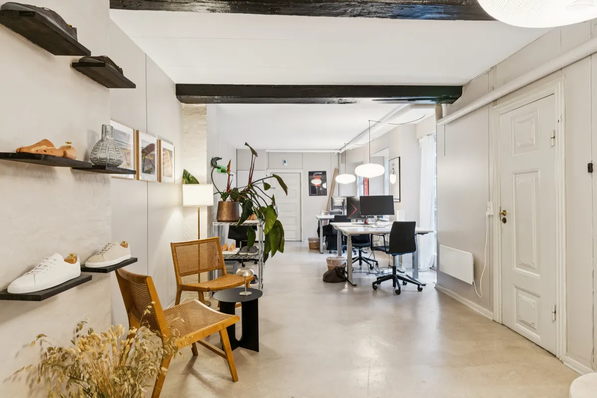 Kontor med hvide vægge, trægulv, højhængende loftsbjælker, tre skohylder med sko, tre stole, to skriveborde, en lampe, en vase, en plante og to stående lamper