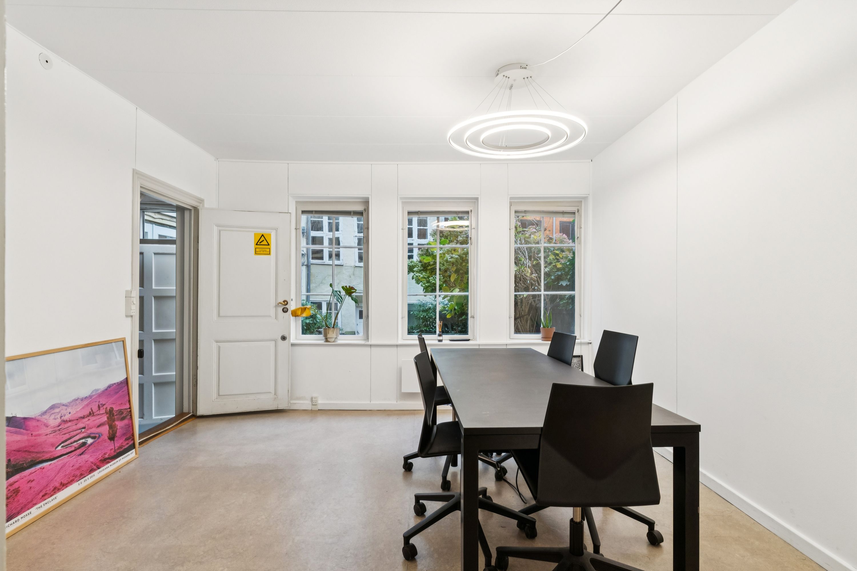 Kontor med højt til loftet, lys, lyse gulve og møbler i sort og hvid. Et lille bord med planter på vindueskarmen og en stor billedramme med landskab.