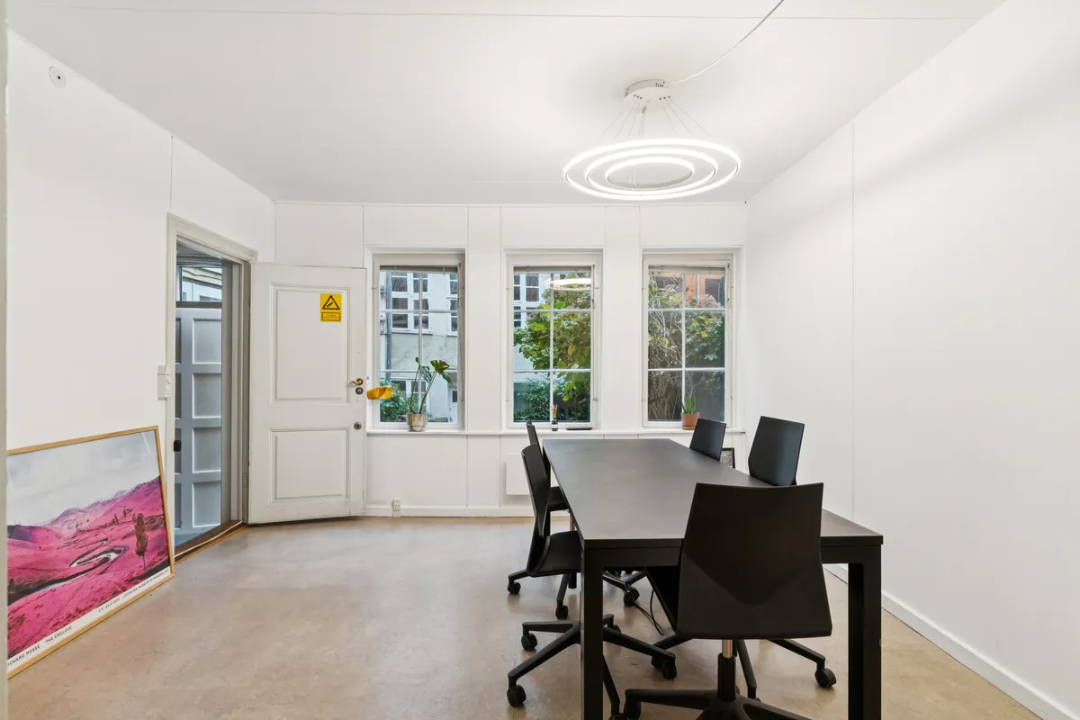 Kontor med højt til loftet, lys, lyse gulve og møbler i sort og hvid. Et lille bord med planter på vindueskarmen og en stor billedramme med landskab.