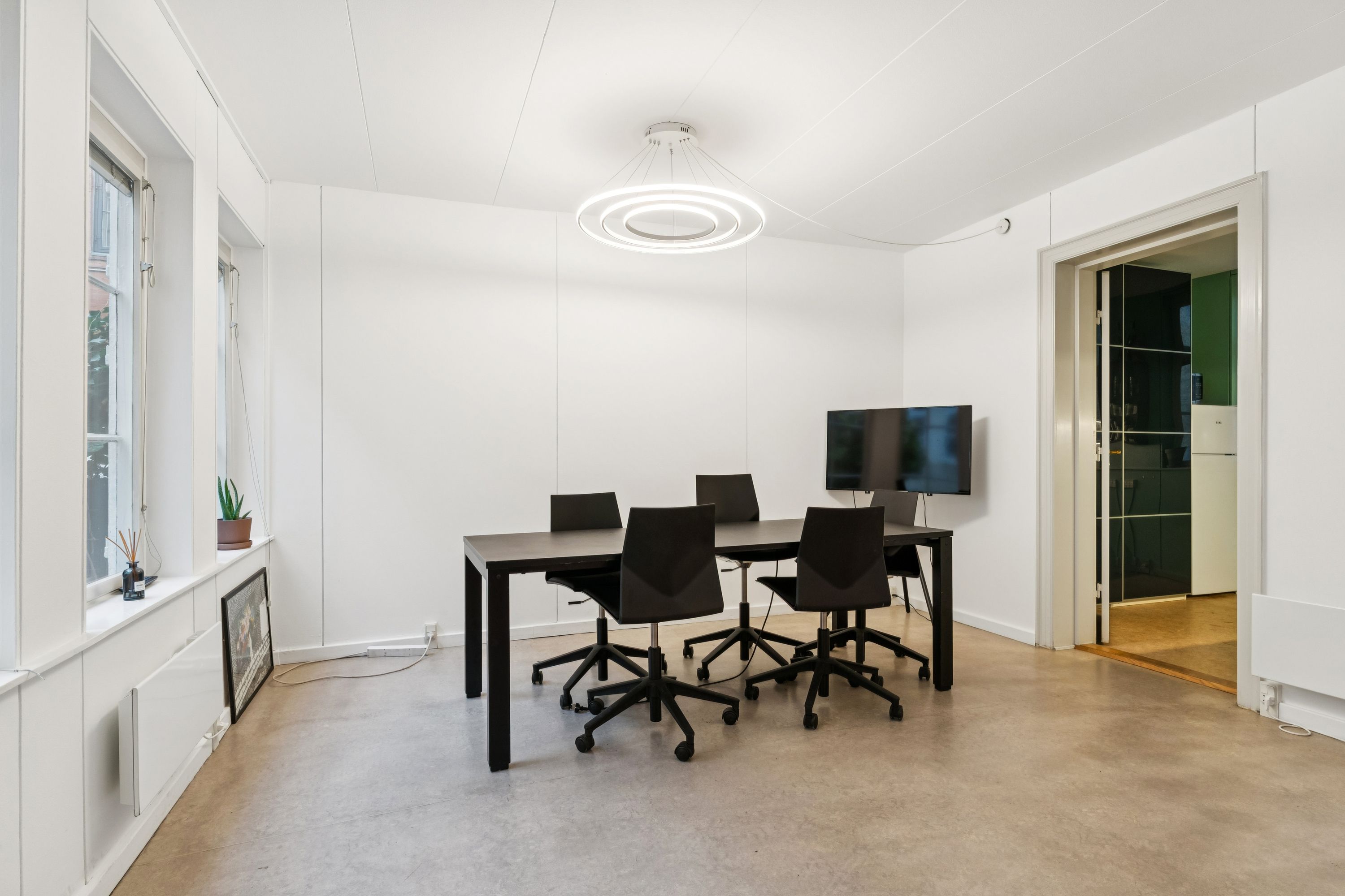 Et lille mødelokale med moderne interiør og minimalistisk design, der inkluderer en rund loftslampe, en stor tv-skærm, et bord og fire stole