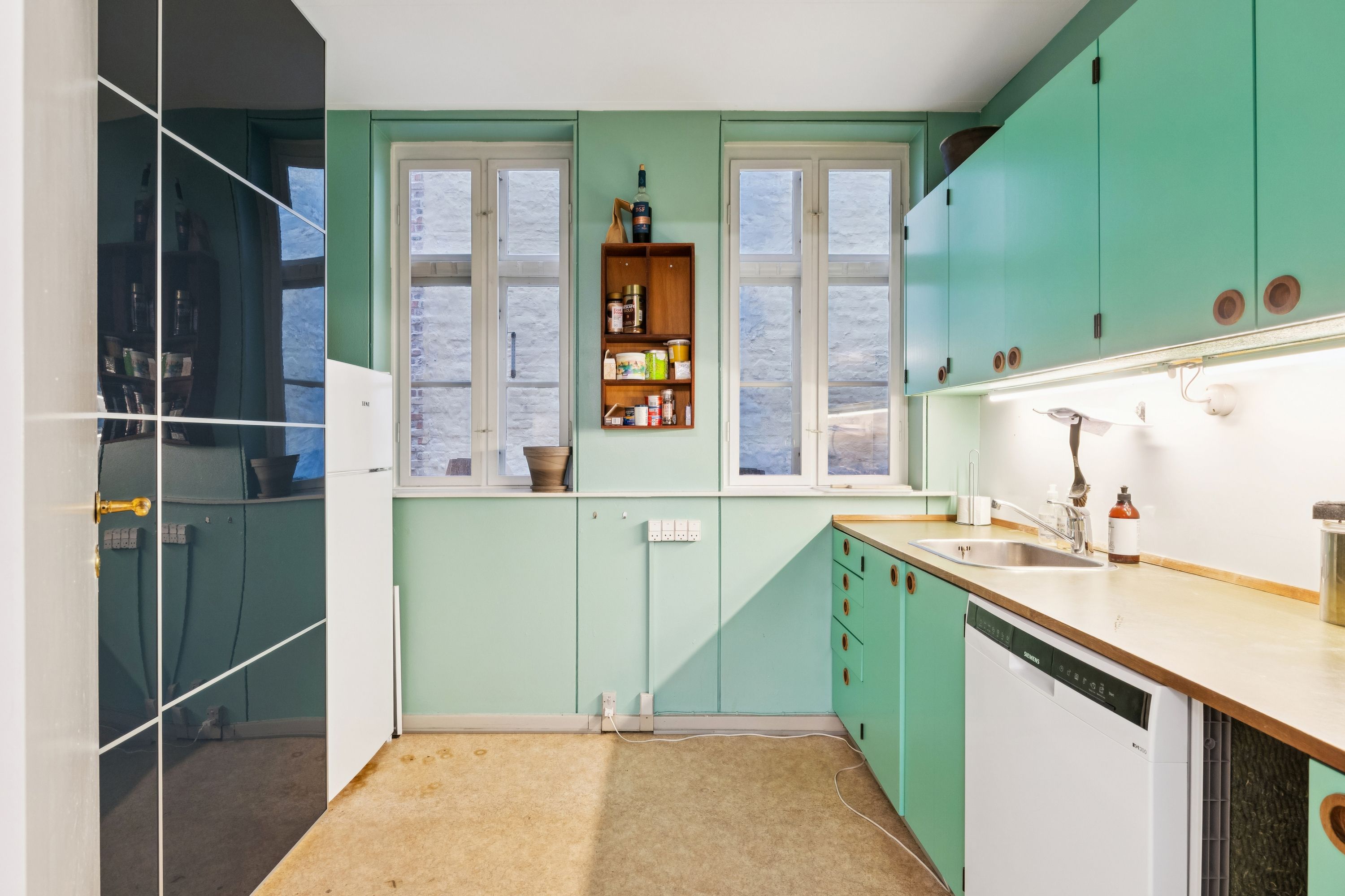 Mintgrønne vægge med hvide ruder og lyse hjørner i køkken med sink og skraldspand. Køkken med hvide apparater og grønne skabe og en åben dør til et moderne badeværelse