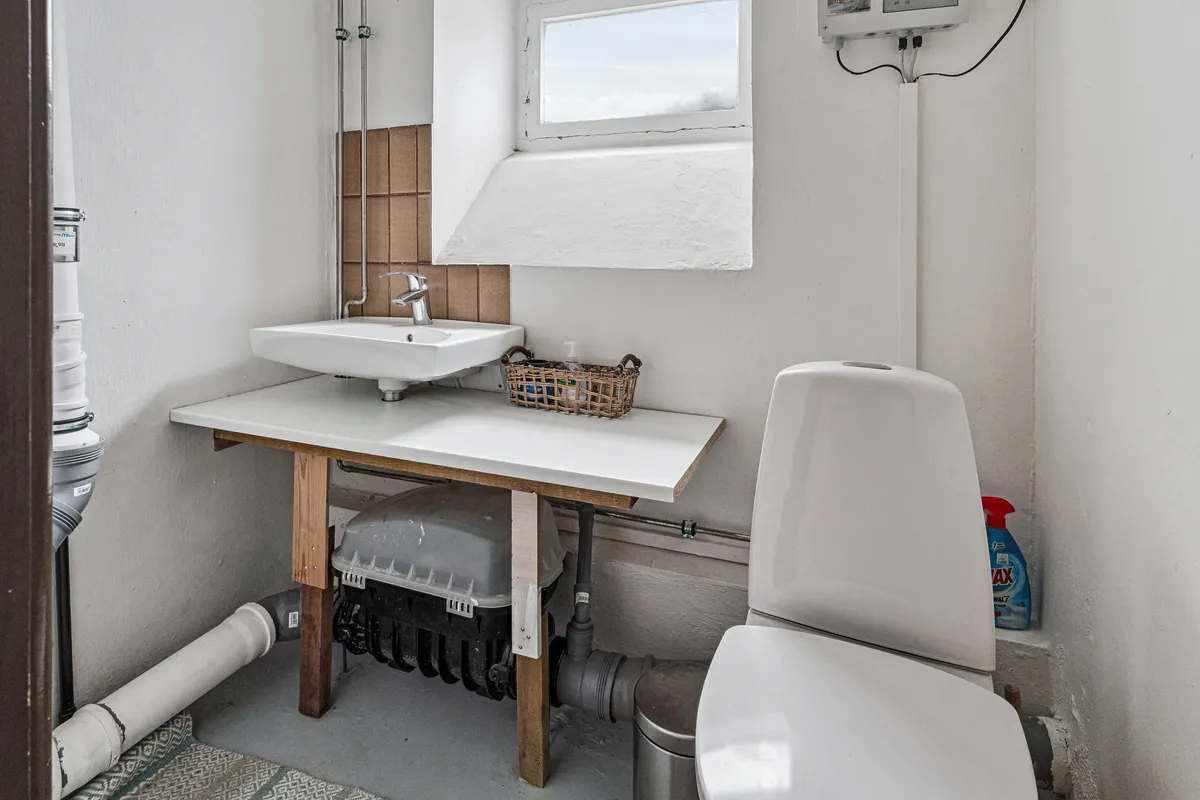 Rummelig badeværelse med hvidt toilet, undertøjsskab, hvidt vasketøjsbord og lysgrå varmeapparat i loftet med badeværelsesventilator