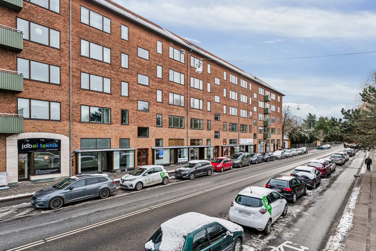 En række parkerede biler på en vej med sne ved siden af en stor bygning med røde mursten og mange vinduer