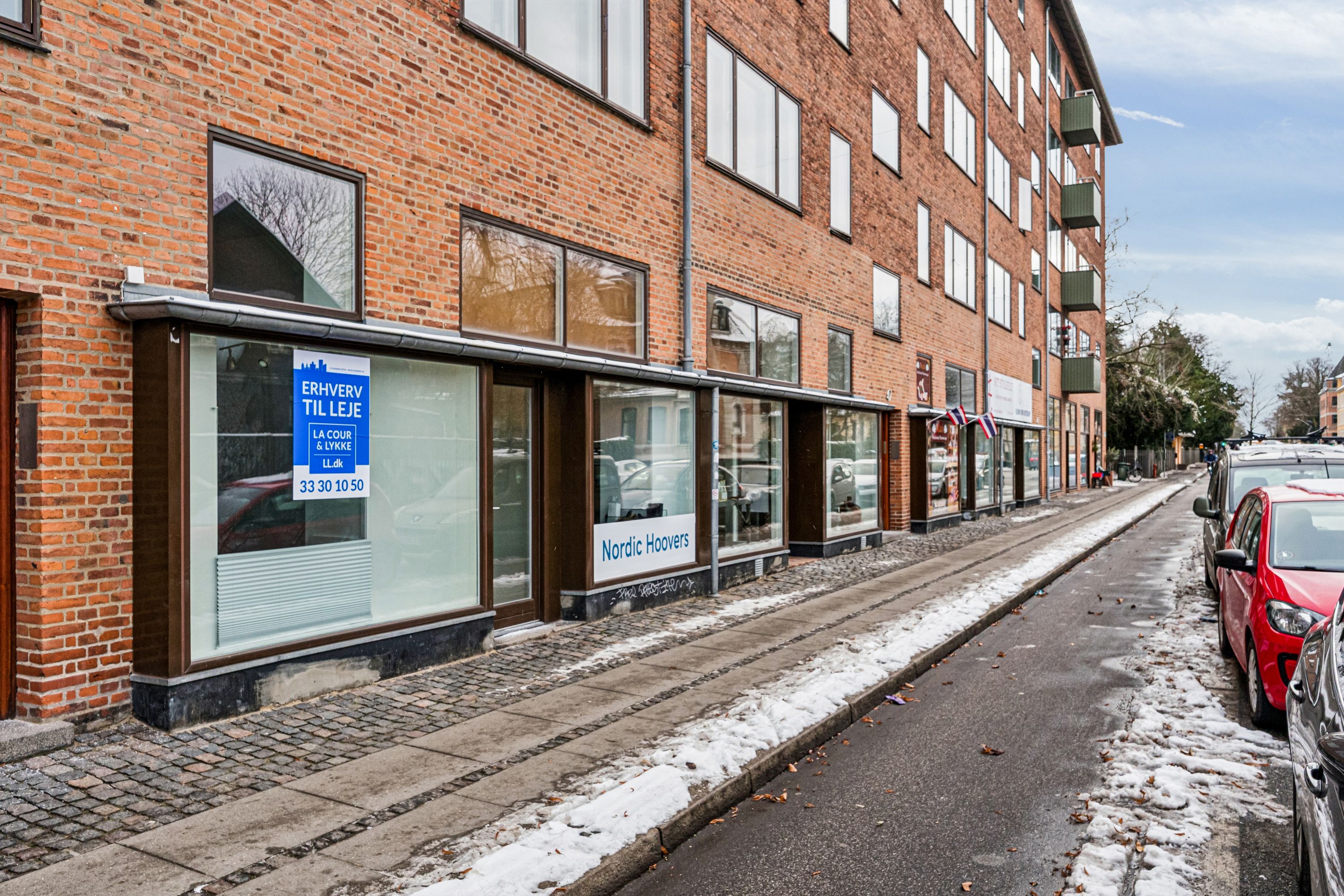 Butik med skilt med teksten Nordic Hoovers til leje på Søndre Fasanvej 45, Frederiksberg. Adgangsvej med sne og biler parkeret langs siden. Vindue med skilt med teksten Erhverv til leje.