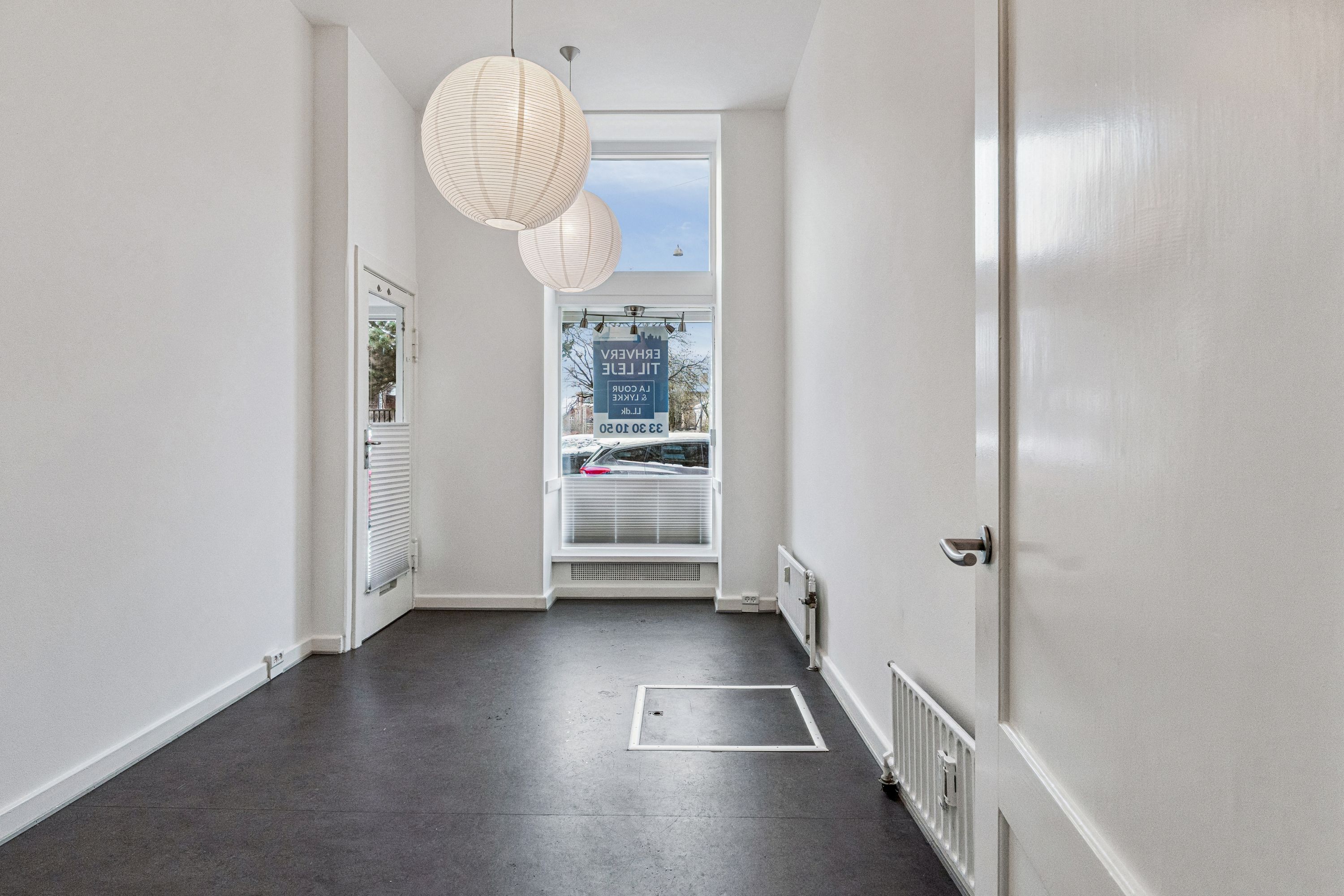 Rummelig og lys indgangshal til butik med højloft og vinduer. Hvide vægge, lyse lysekroner og rummelige gulve. Lejemålet består af stueplan og et baglokale.