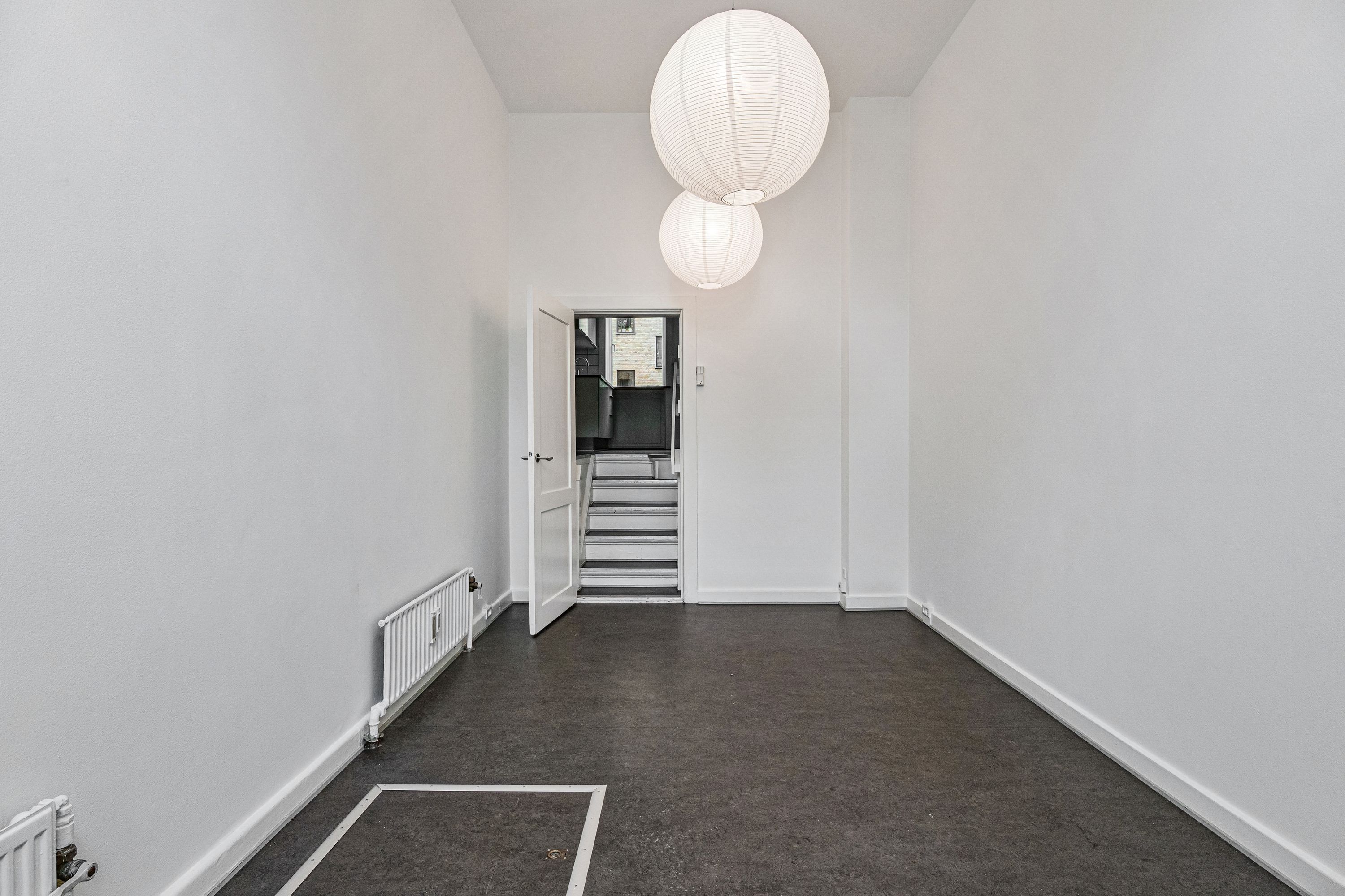 En stor stue med højloft og åben dør til gården. To hvide kuglepærer hængende på loftet og en hvid radiator på højre side