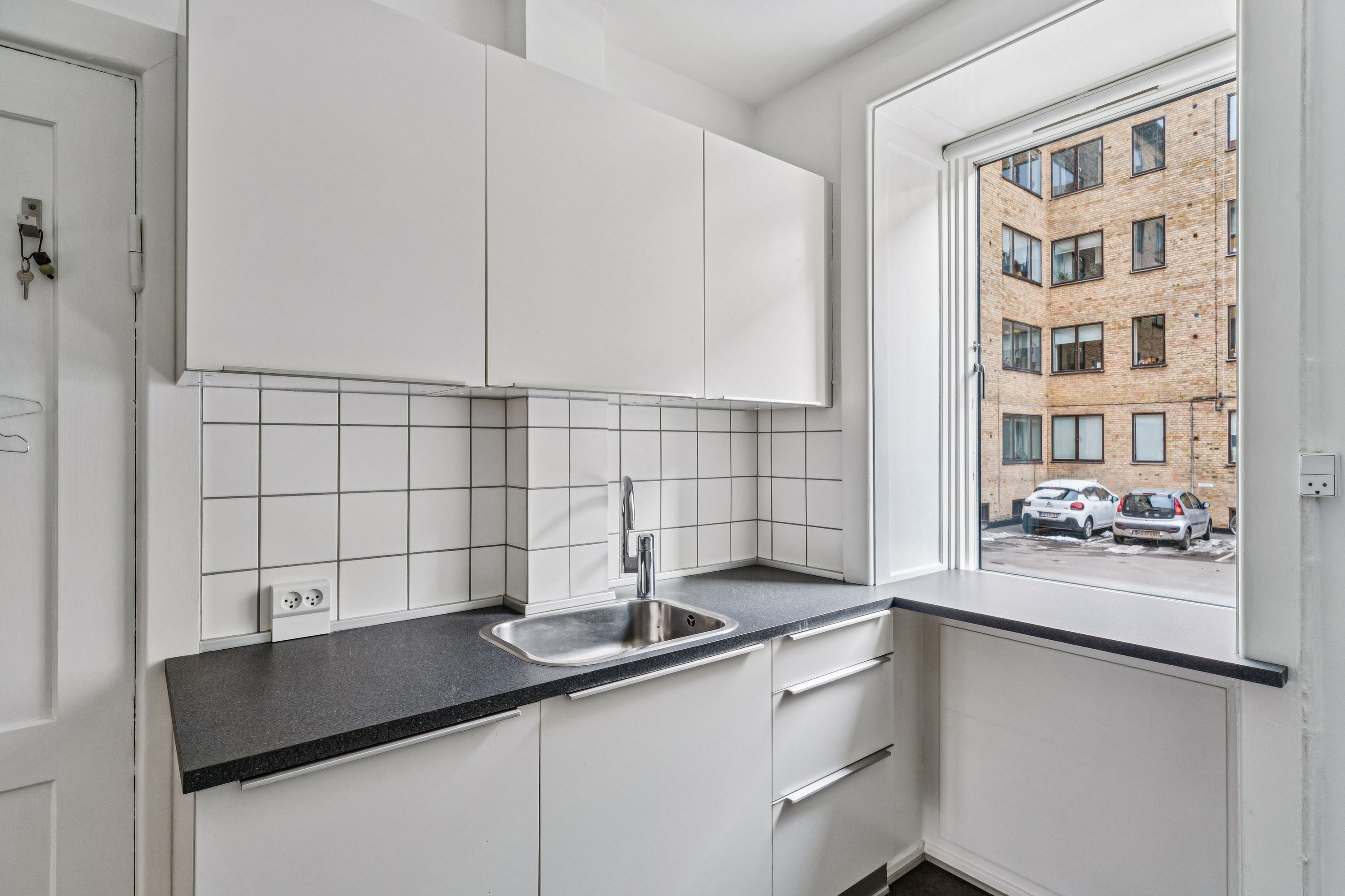 Hvidt køkken med højloftede rum og udsigt til parkeringsplads og lejlighedsbygning, hvor to biler er parkeret.