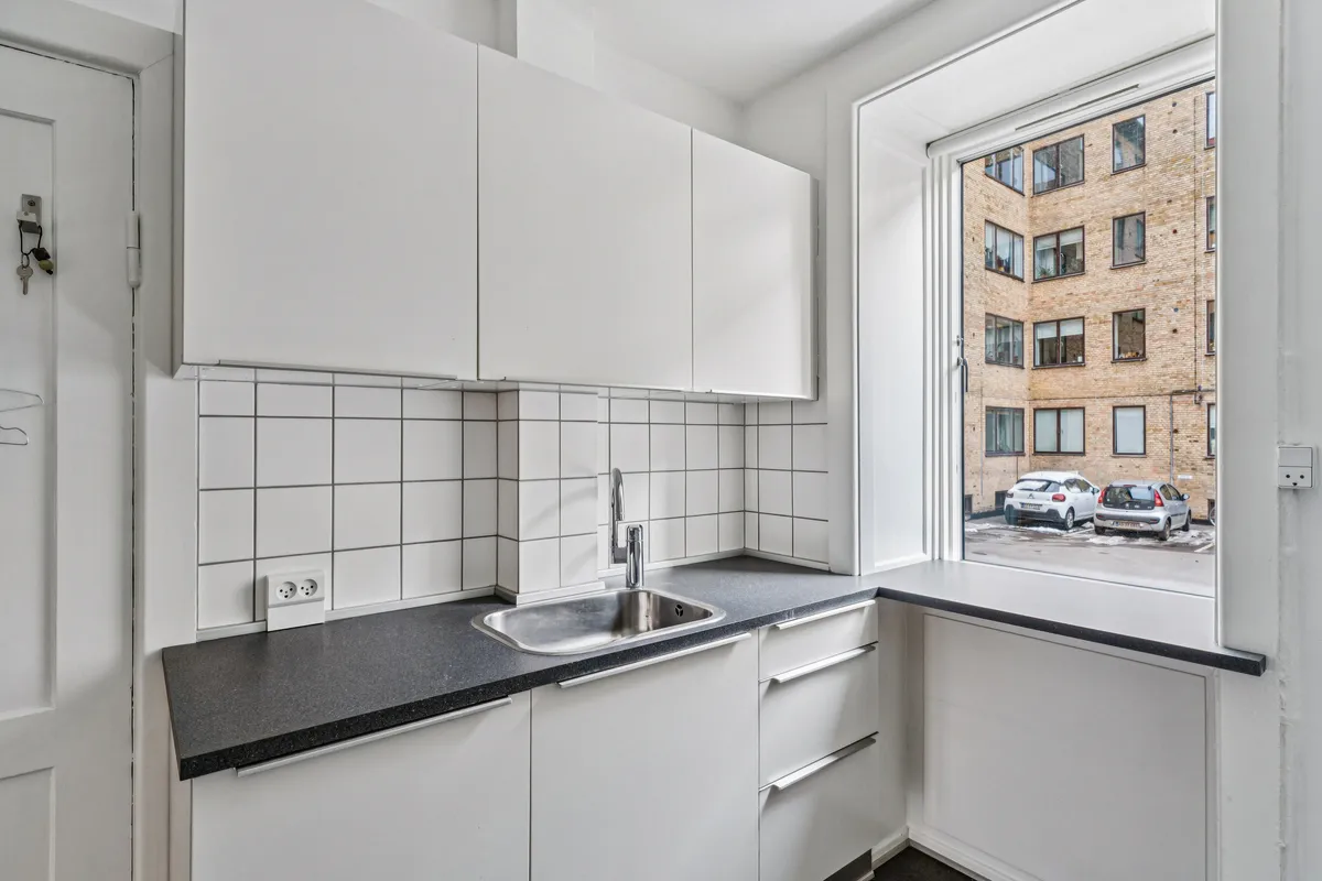 Hvidt køkken med højloftede rum og udsigt til parkeringsplads og lejlighedsbygning, hvor to biler er parkeret.