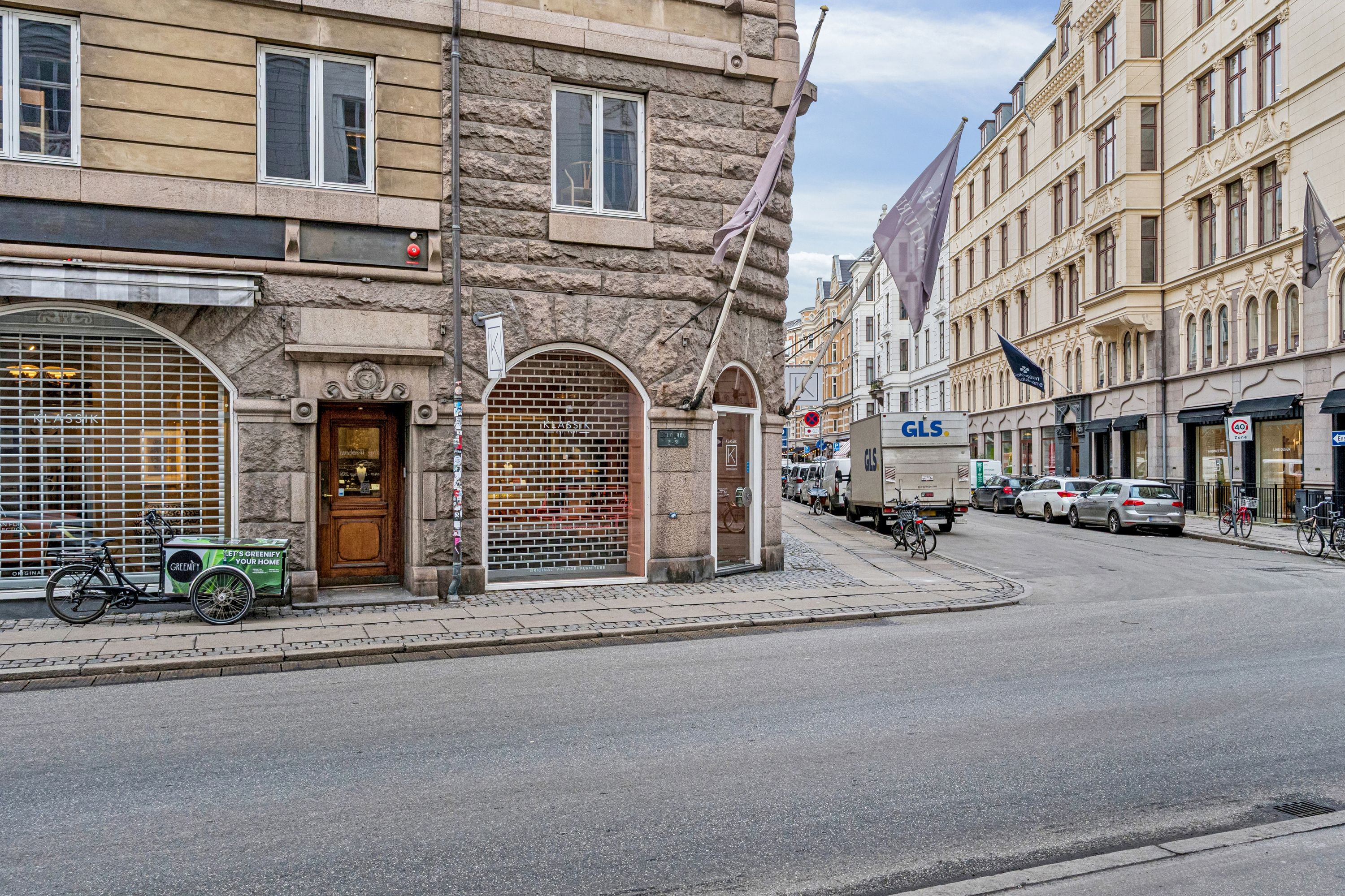 To biler og en lastbil parkeret på Bredgade. To cykler står udenfor forretning. En trehjulet vogn står parkeret foran en dør. Flag hængende på bygningen.