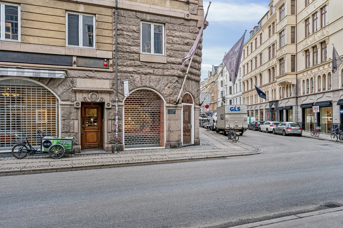 To biler og en lastbil parkeret på Bredgade. To cykler står udenfor forretning. En trehjulet vogn står parkeret foran en dør. Flag hængende på bygningen.