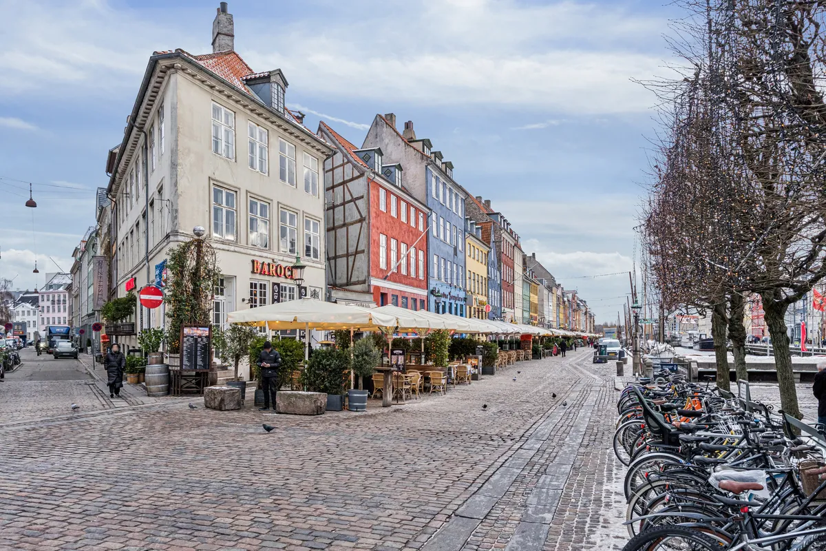 En bred gågade med række af gamle røde og hvide bygninger med terrasser og planter samt række af cykler parkeret på højre side