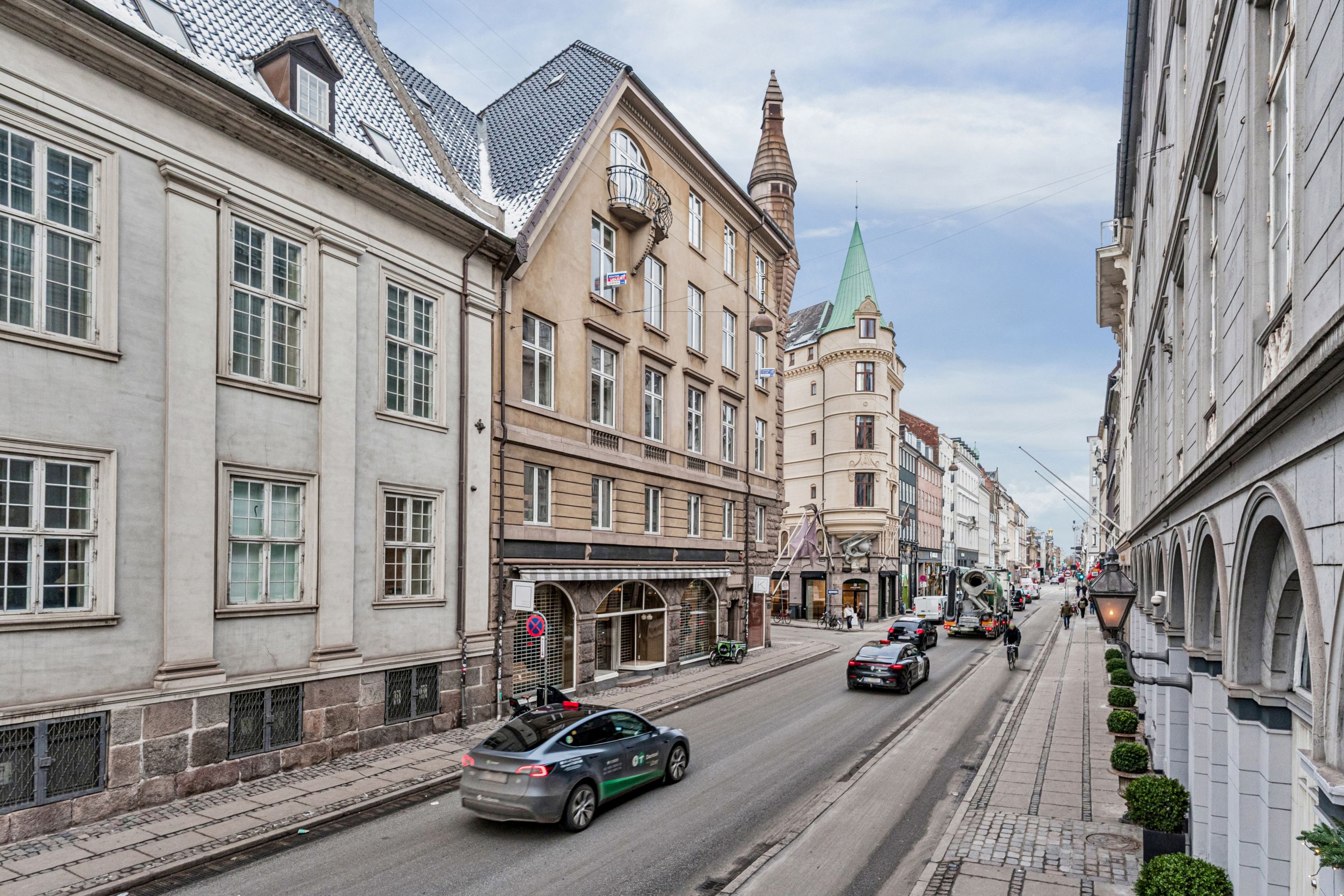 En bil kører på en gade, der er kantede med grønne planter og bygninger med glasruder og søjler