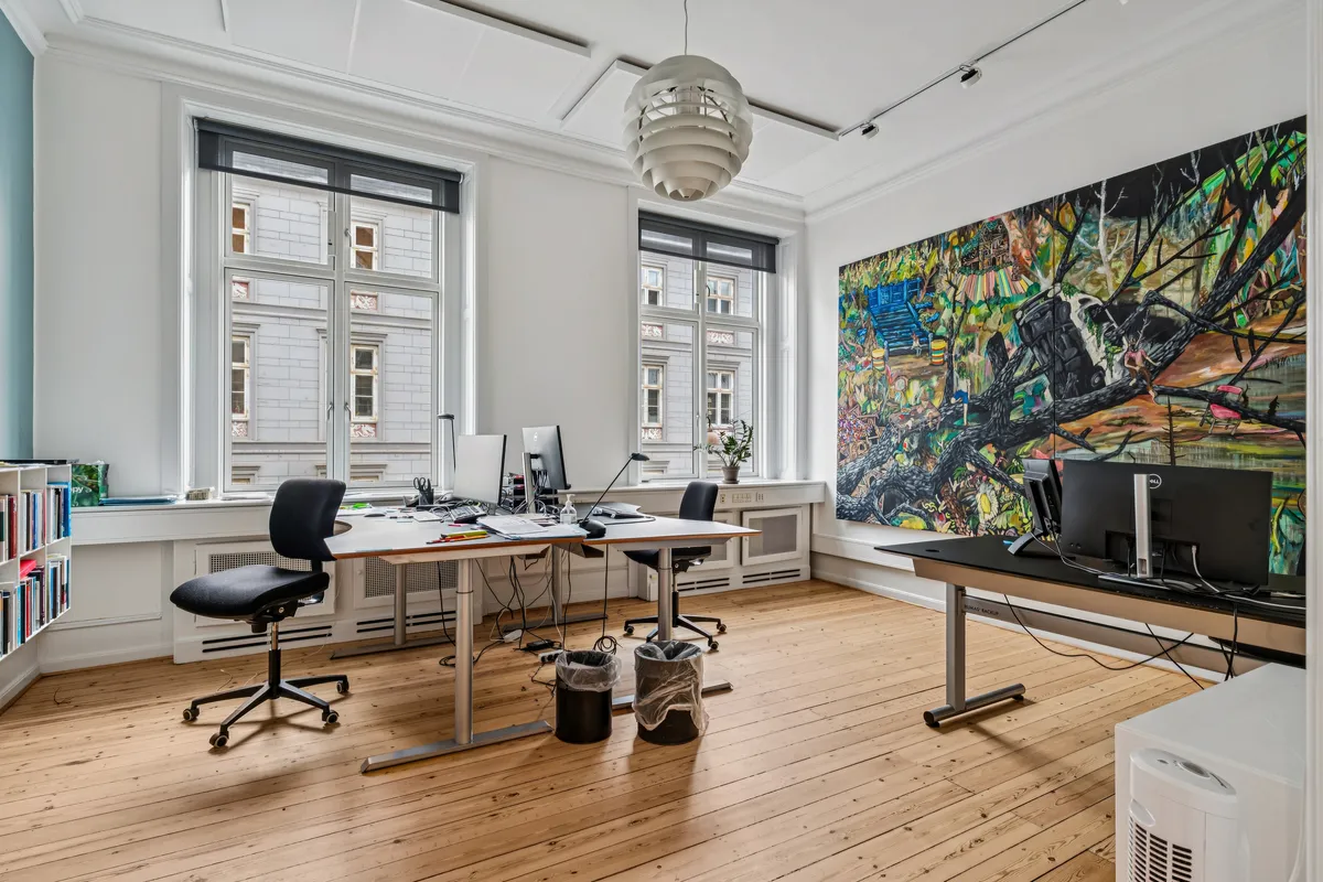Kontor med lyse lofter, vinduer og trægulv. To skriveborde med computere og stolpe til møder. Et stort maleri hænger på væggen