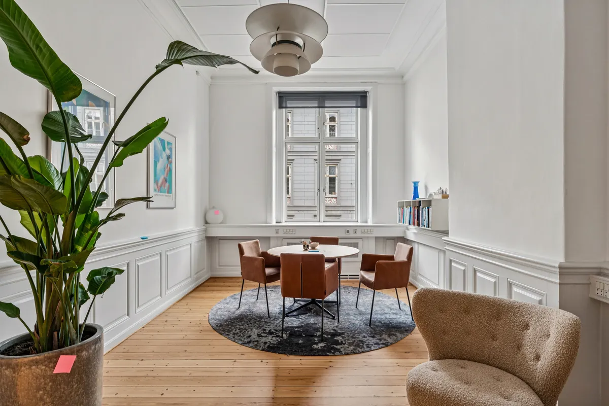 Kontorrum med moderne møbler og en stor plante. Bord og stole samt en sofistikeret lampe. Vindue med udsigt til en bygning og lyse skyer. En hylde med bøger og en dekorativ vase