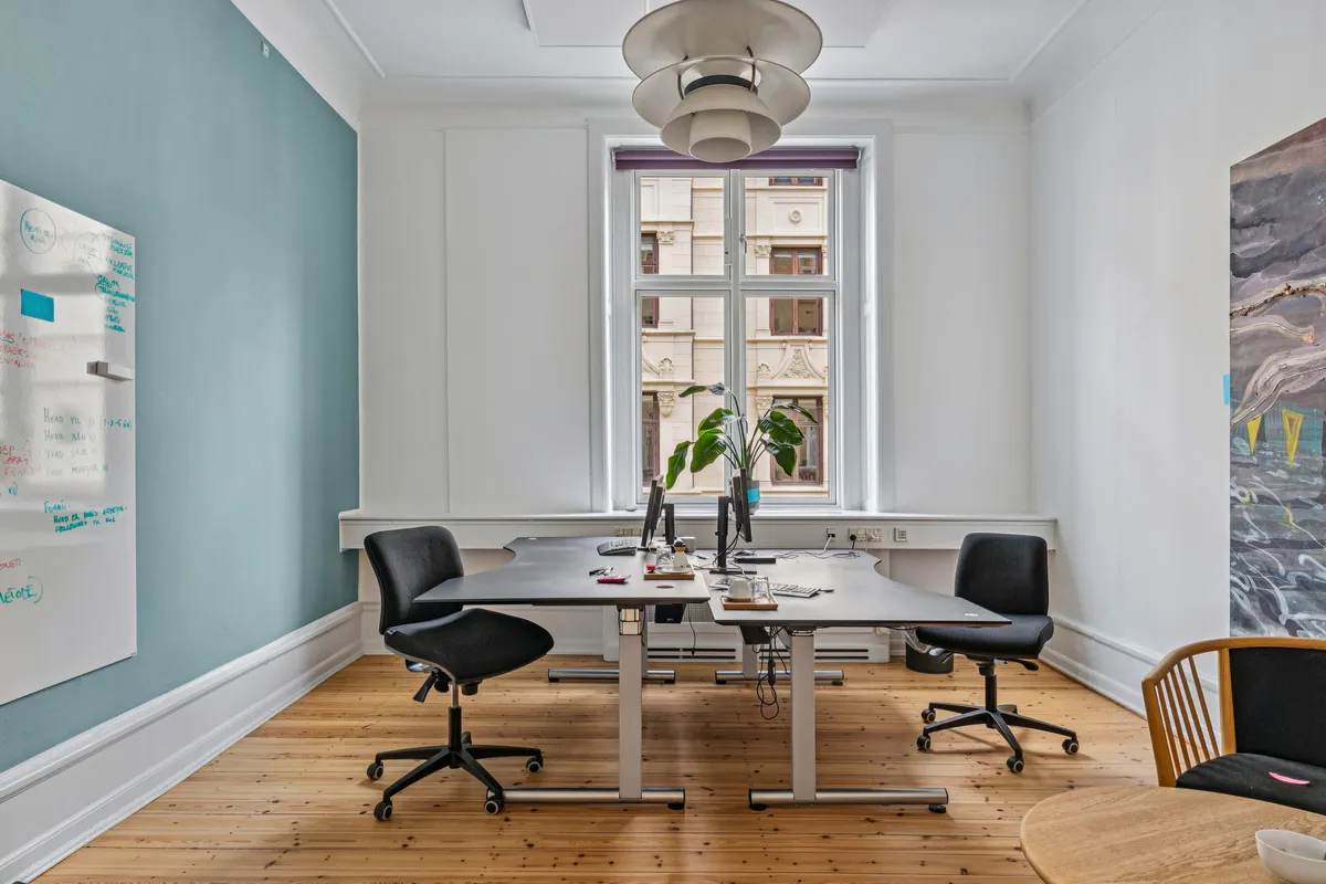 Kontorrum med bord og skrivebord, skrivebordslampe og en plante i vinduet samt et tavle og en stol ved et mindre rundt bord