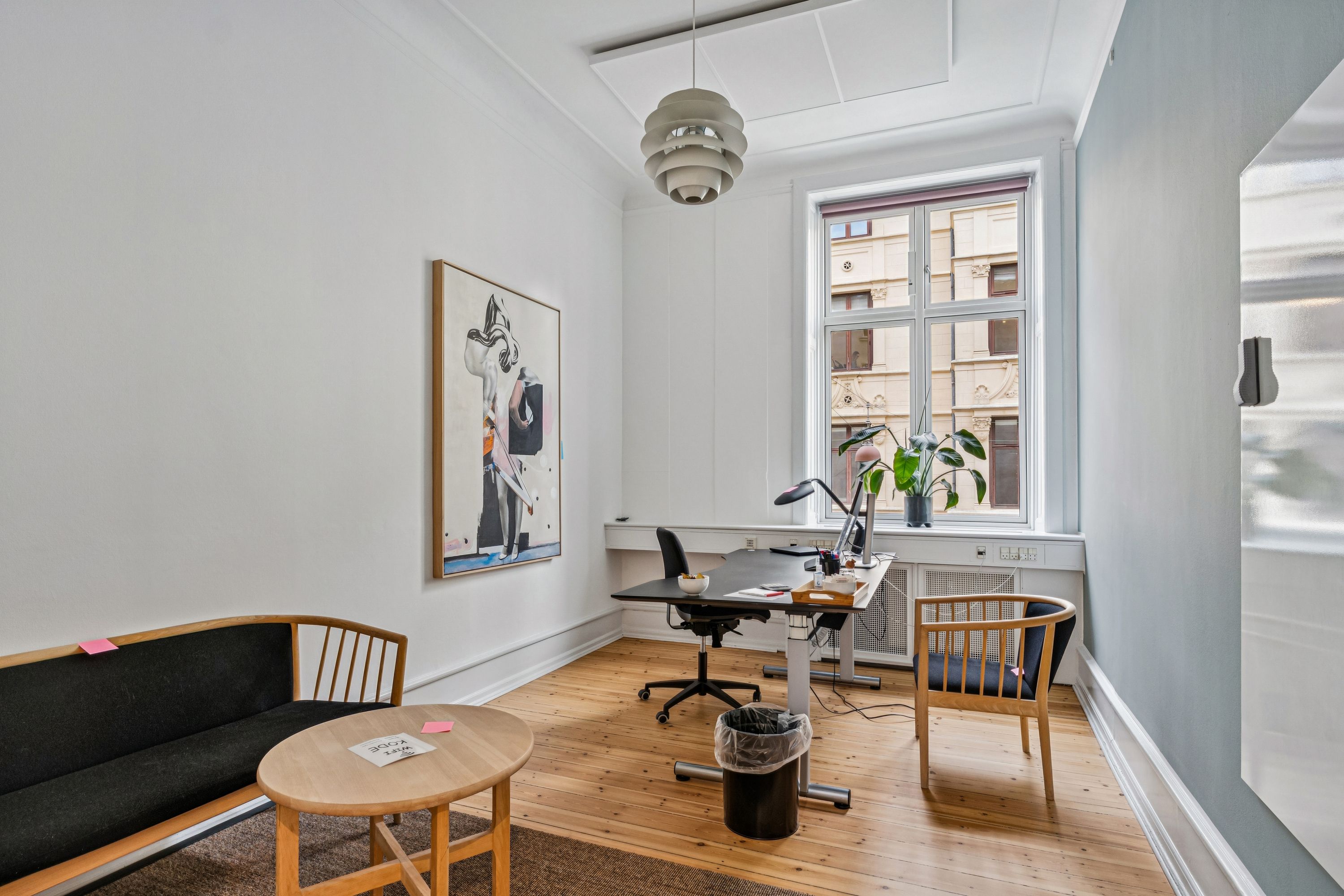 Kontor med sofa, skrivebord, stol, plante, kunstværk og vinduesvisning til bygning og vej