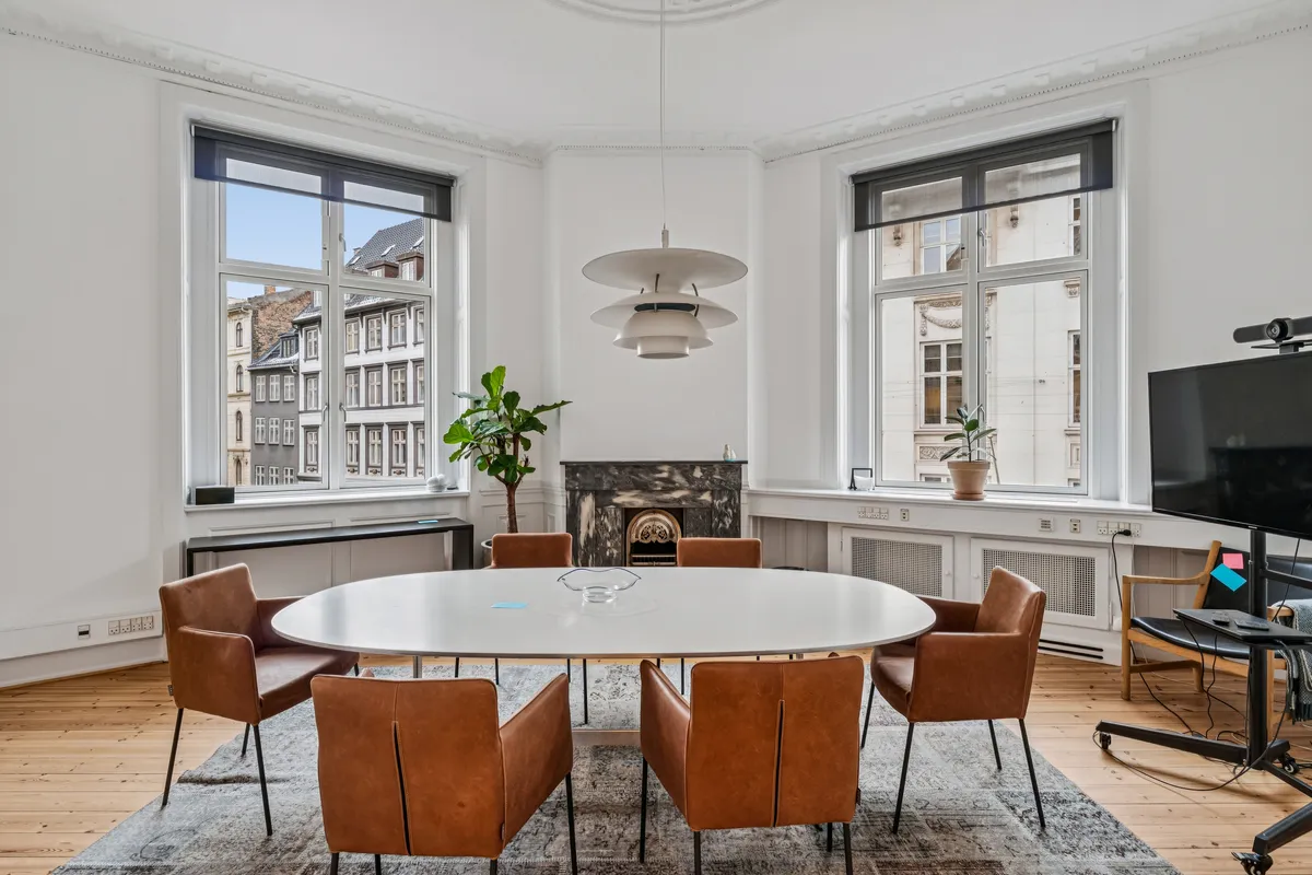 Rundt kontor med runde borde og stole med læderopslag og lysekroner, to plantepoter og tv. Vinduesglas med gardiner og en skorsten i baggrunden.