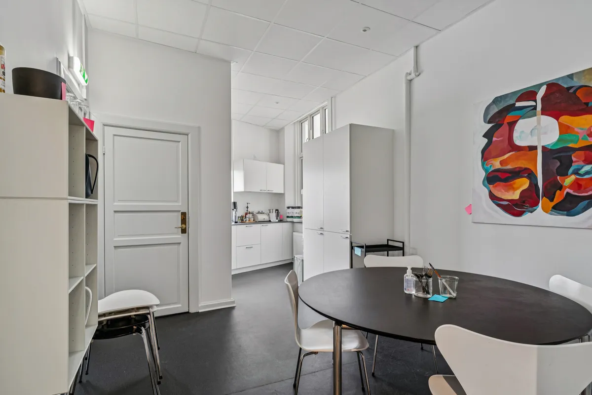 Rundt bord med fem stole og stol med lyserøde stifter i minimalistisk kontor med hvide vægge og loft. Farverige malerier og høj loftshøjde.