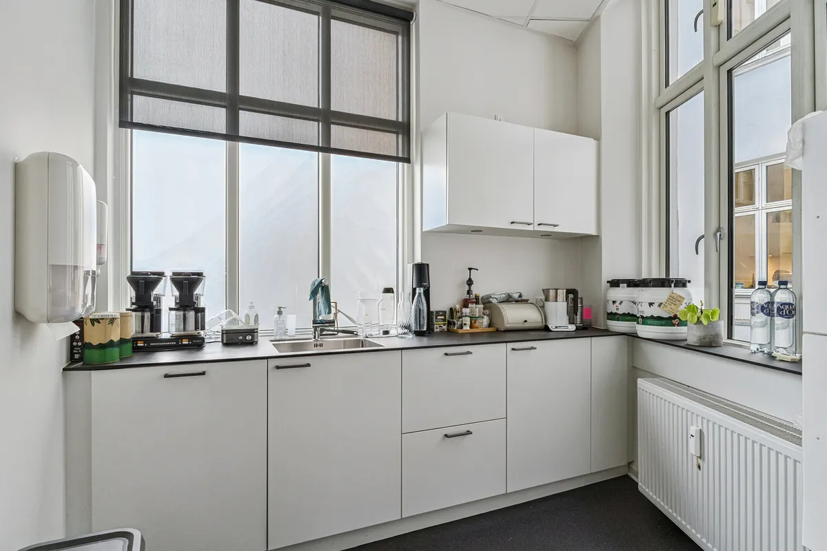 Kontor til leje på Bredgade 3, 2., 1260 København K med moderne køkken med hvide køkkenskabe, sink, gryder og apparater. Køkkenet har store vinduer og et vindue med udgang til gaden.