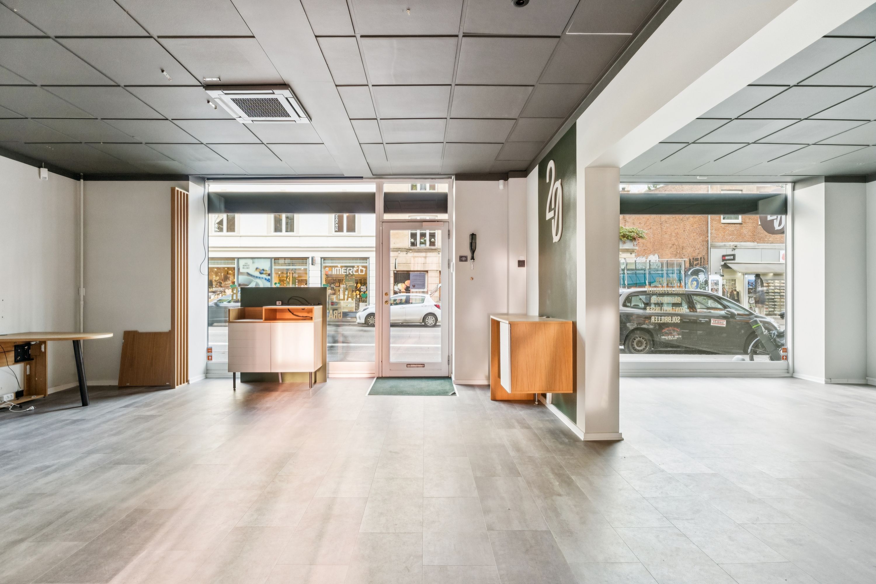 Indersiden af en tom butik med lyse gulvfliser og hvide vægge, med udsigt til en glasdør og vej, hvor biler kører forbi