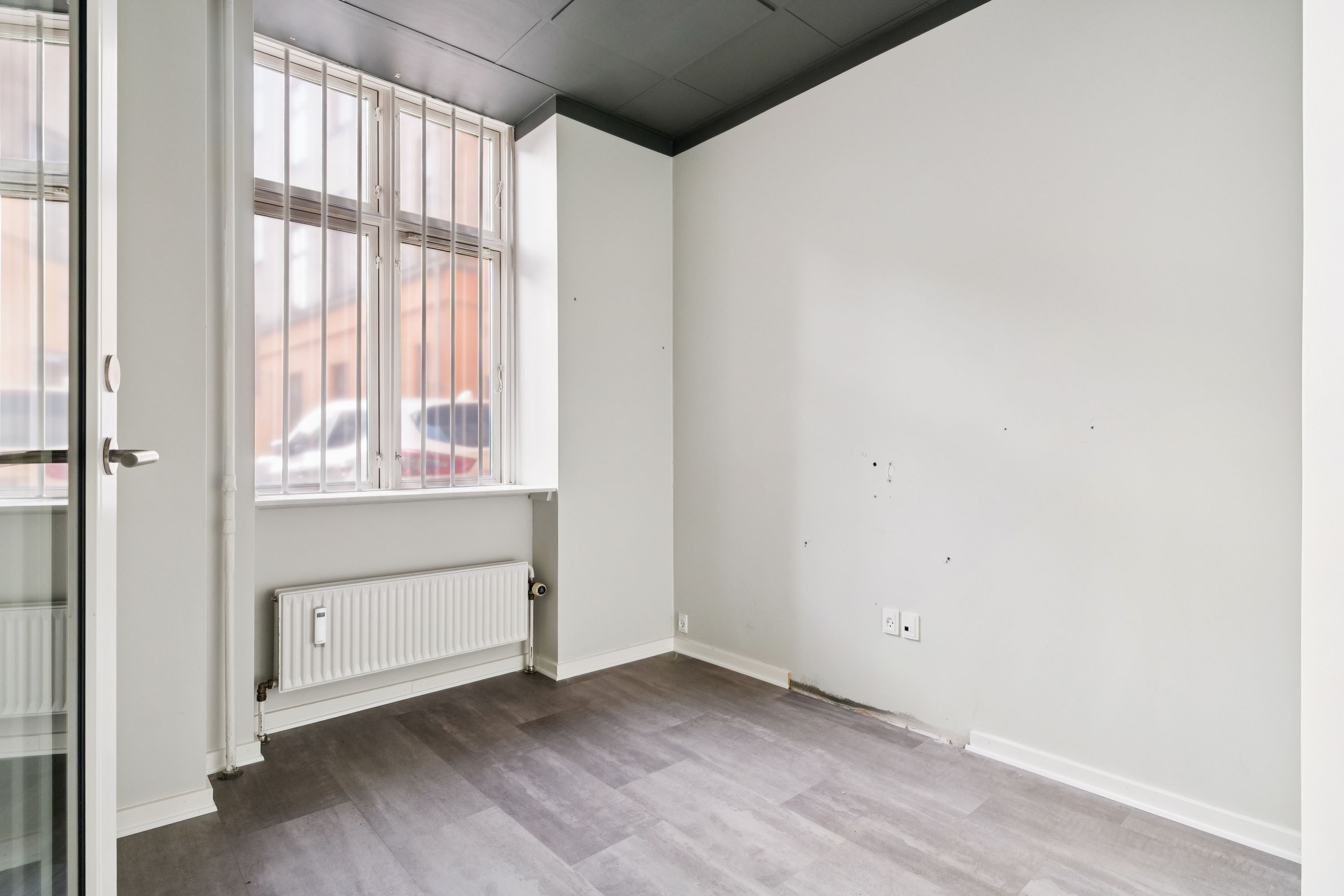 En stor butikslokale med lyse gulve, en stor rude og et radiator under vinduet samt et dørhåndtag og en lukket glasdør