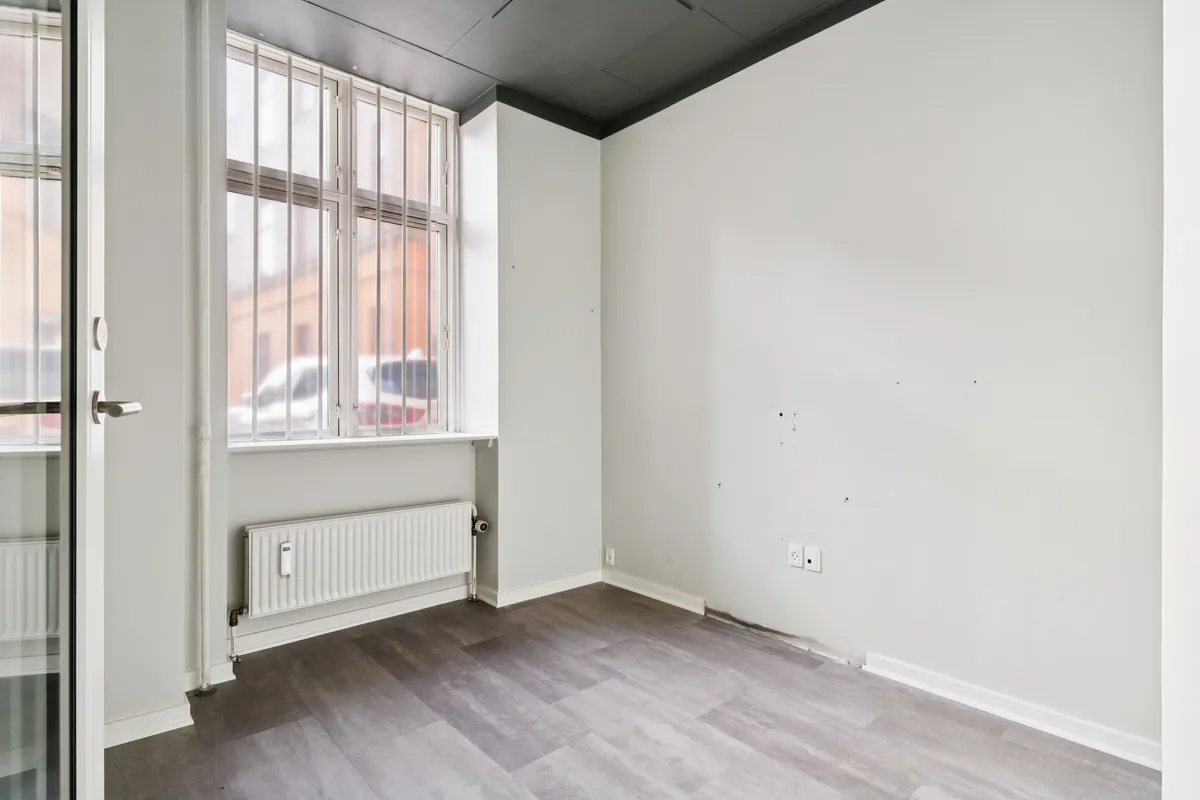 En stor butikslokale med lyse gulve, en stor rude og et radiator under vinduet samt et dørhåndtag og en lukket glasdør