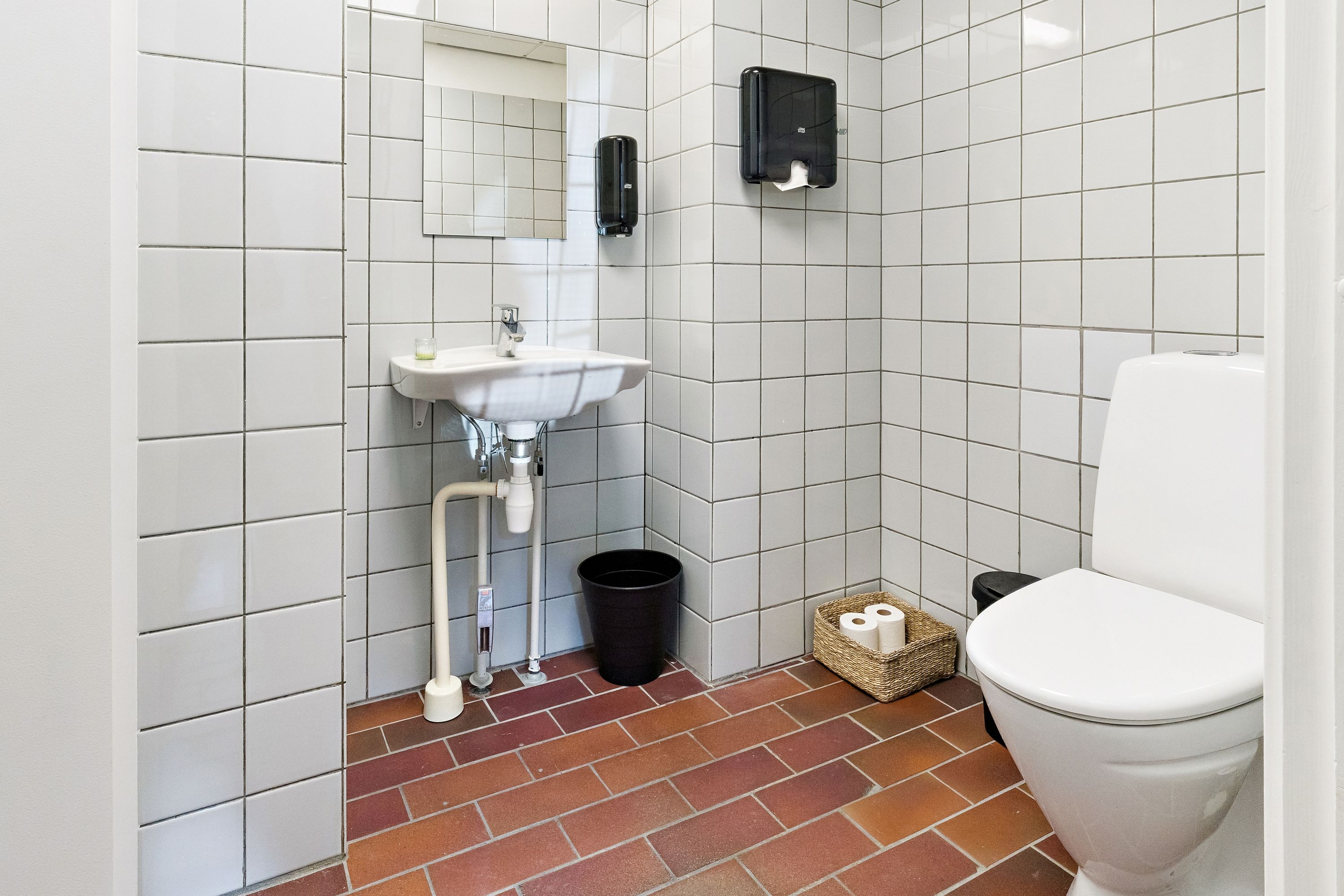 En lille badeværelse med hvide fliser, en hvid vask med en håndfat, to håndklædeholdere og et toilet. Et toiletpapirudstyr og en papirkurv er placeret på gulvet.